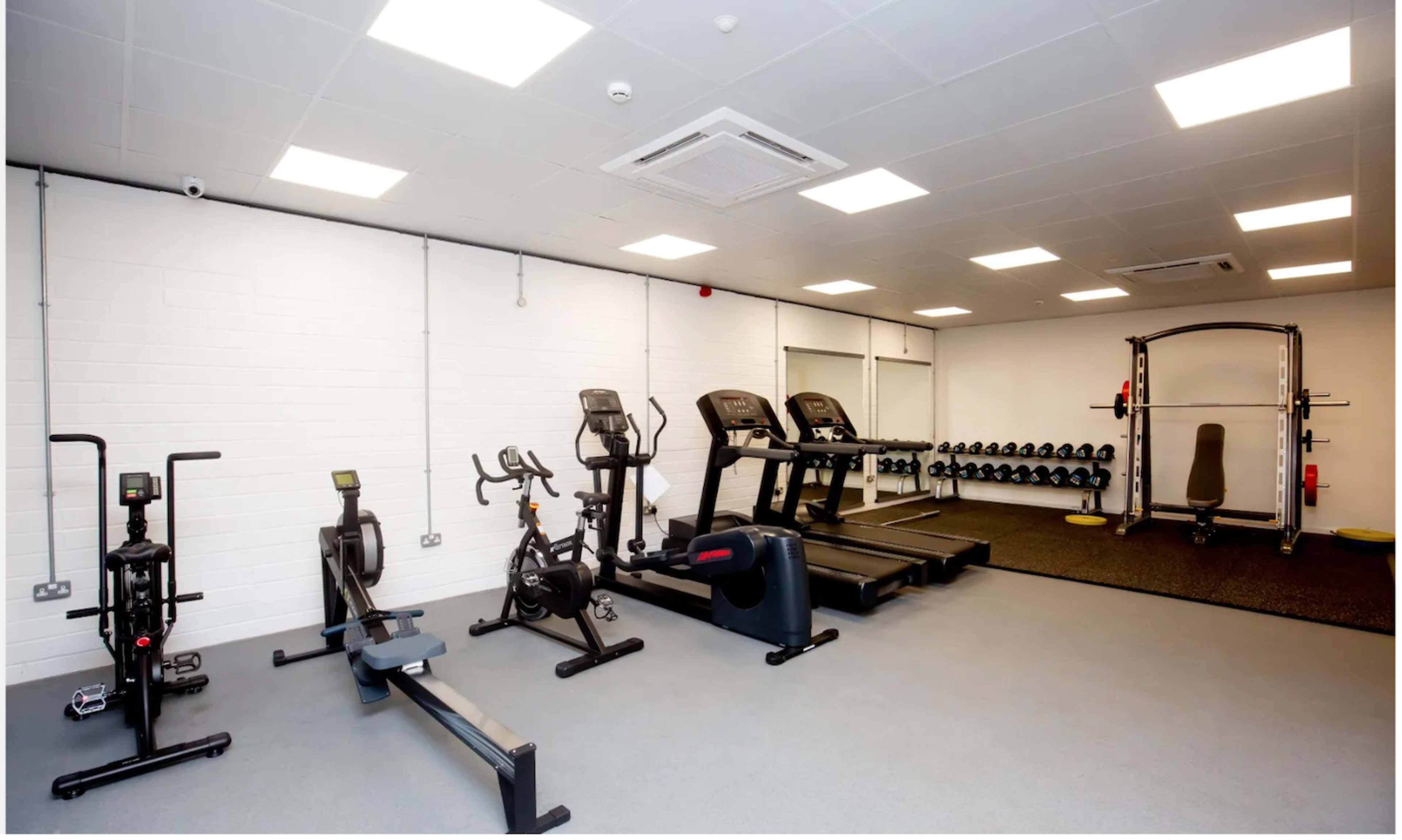The image shows an indoor gym with various exercise equipment, including treadmills, a rowing machine, a stationary bike, free weights, and a multi-gym station.