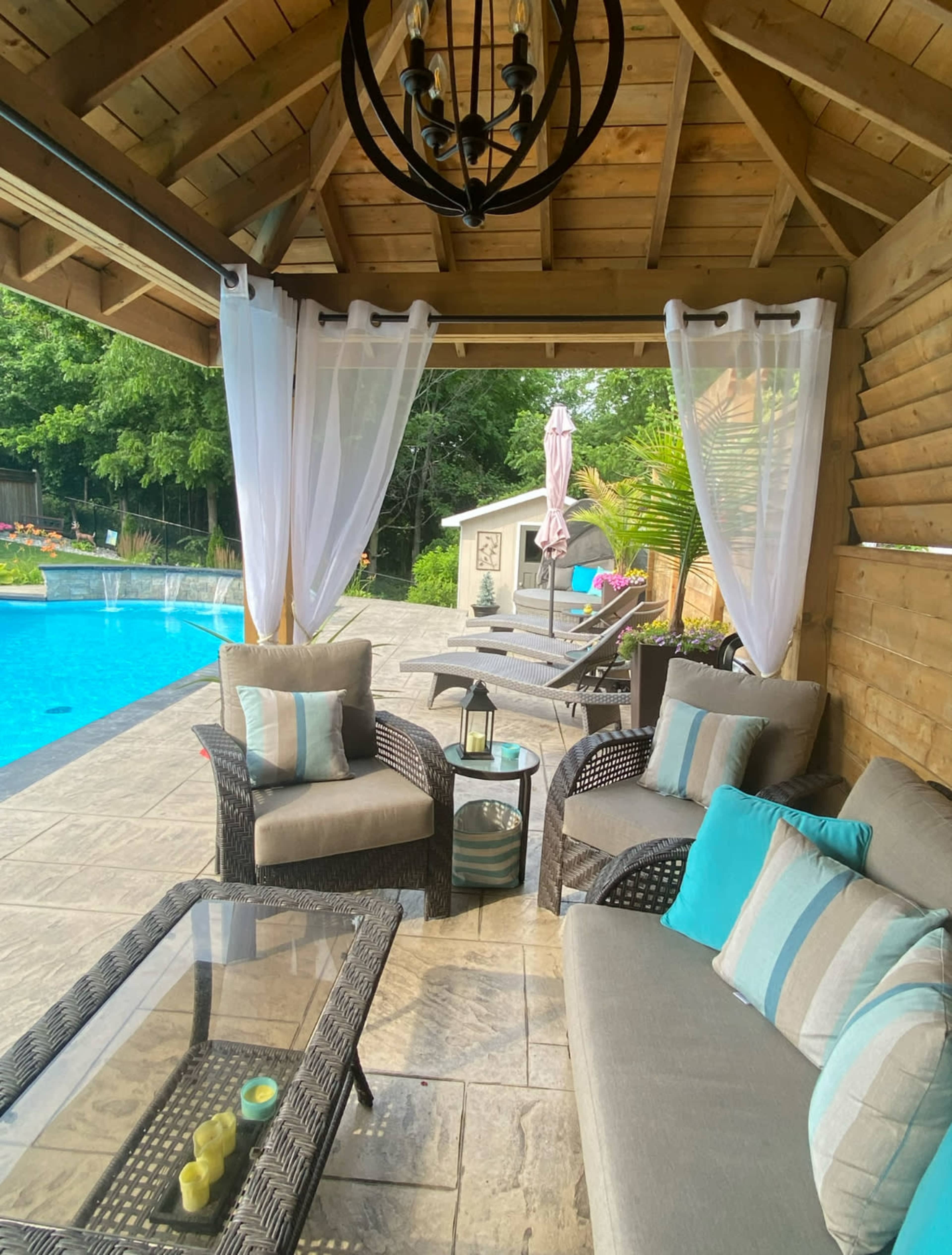 A seating area with wicker furniture and a glass table is situated beside a blue swimming pool, under a wooden gazebo with sheer curtains.