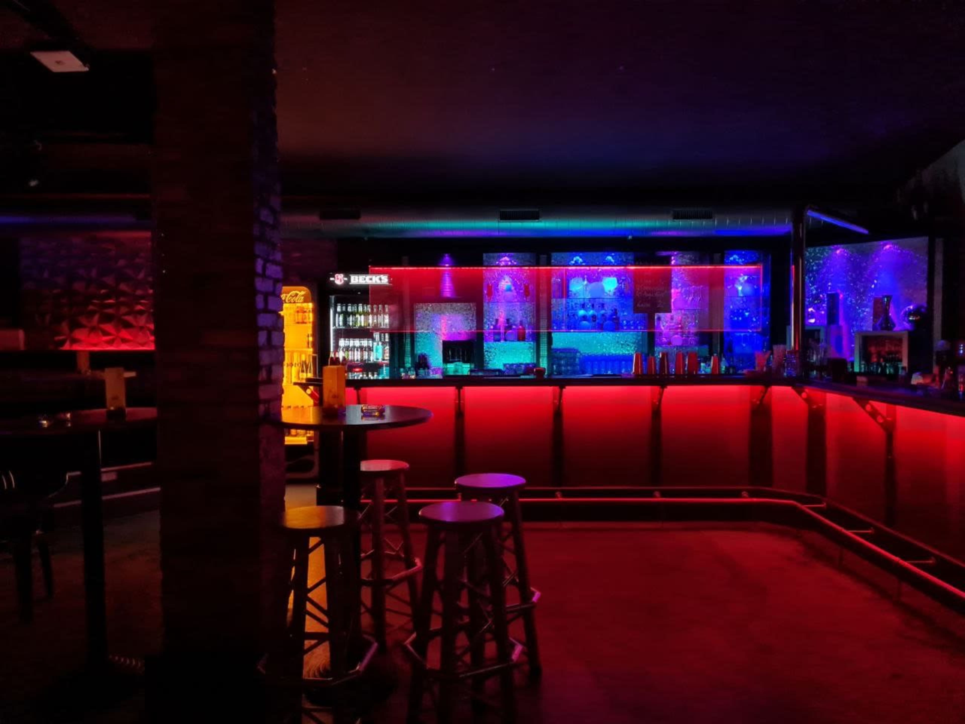 A dimly lit bar with colorful LED lighting highlighting the shelves and counter, featuring high stools and a brick column.