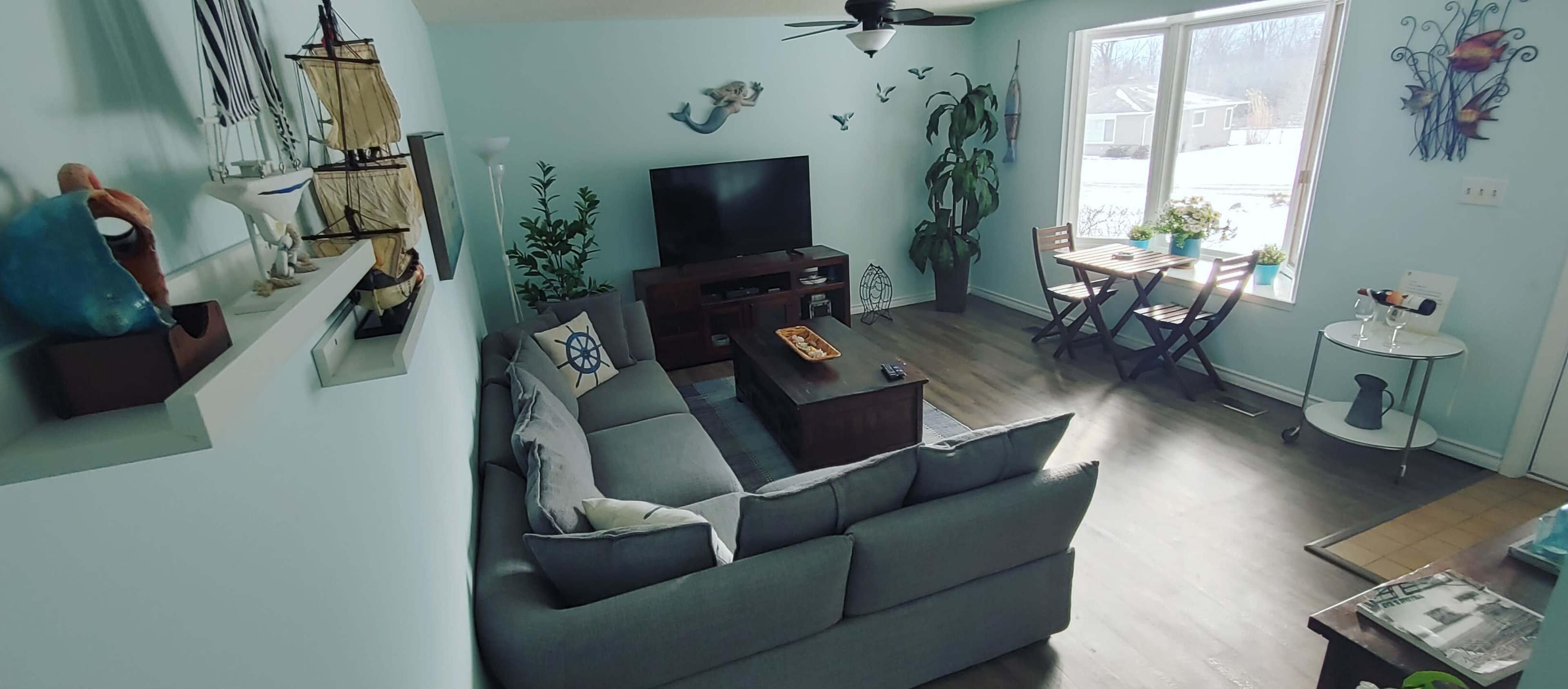The image shows a living room with a gray couch, a wooden coffee table, a television, and a small dining area with a table and chairs by a large window.
