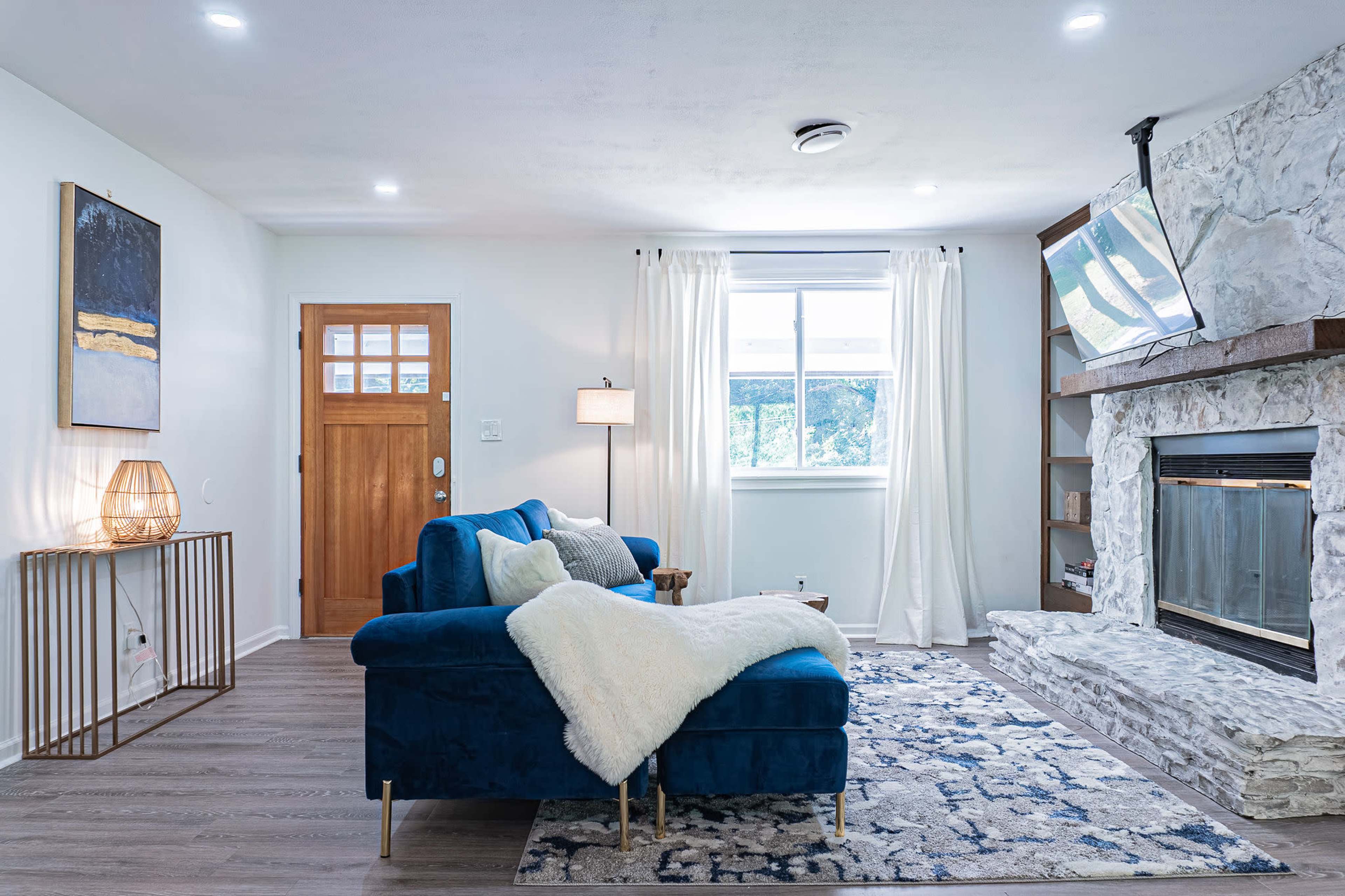 A living room with a blue sectional sofa, a stone fireplace, natural light coming through a window, and a wooden door.