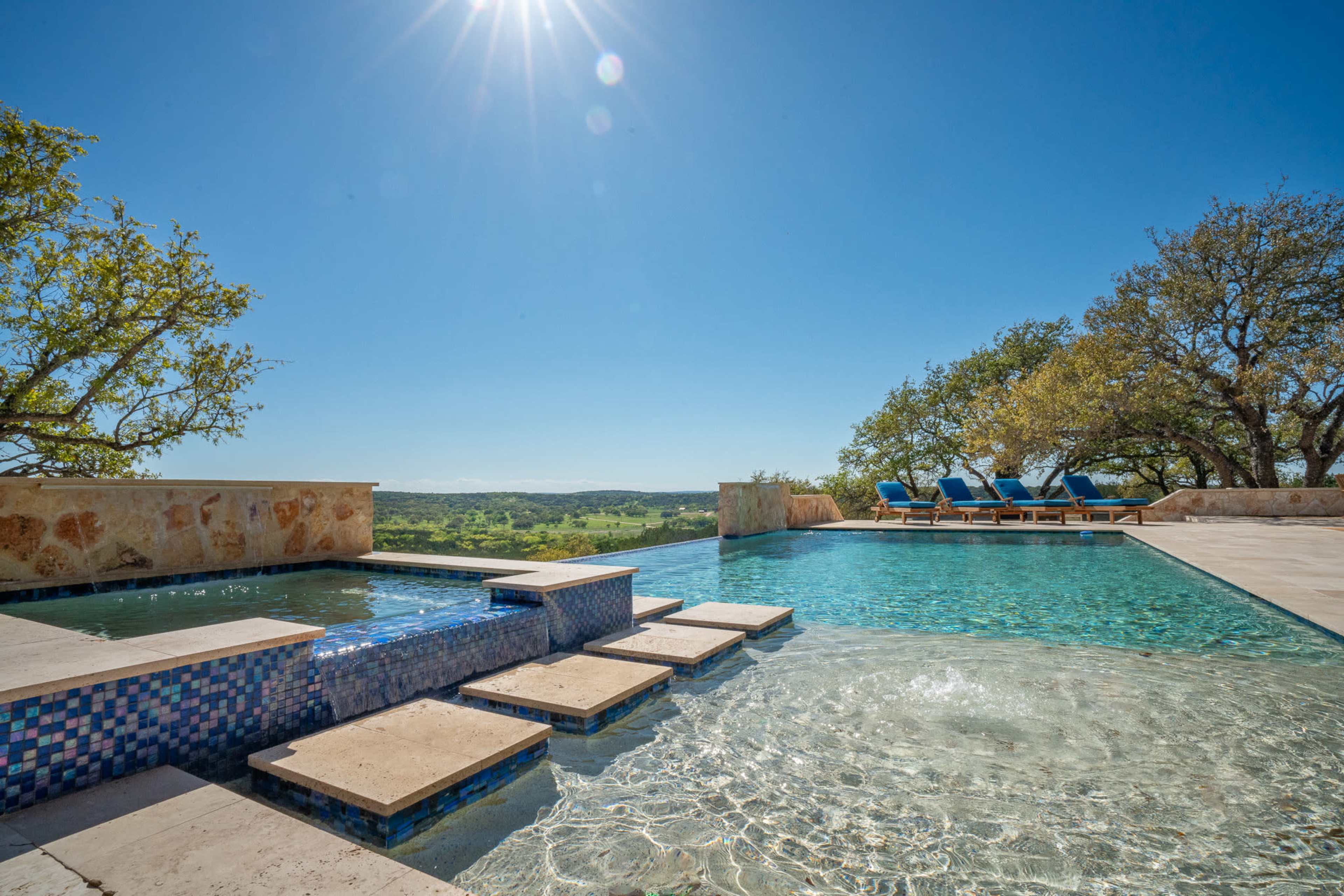 A modern infinity pool with a hot tub and stone steps overlooks a scenic landscape under a clear blue sky.