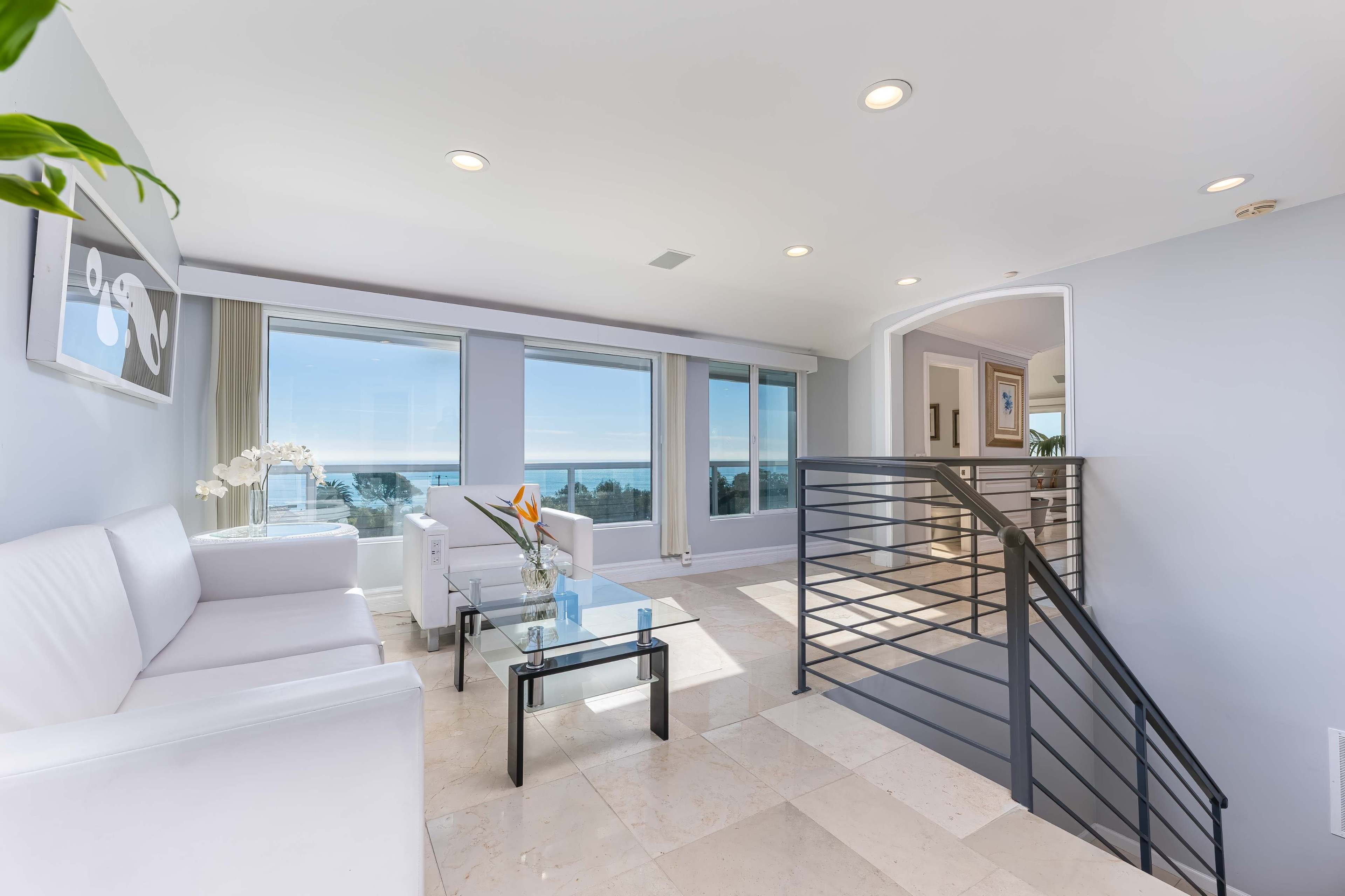 A modern living room with white furniture, a glass coffee table, and large windows offering a view of the ocean.