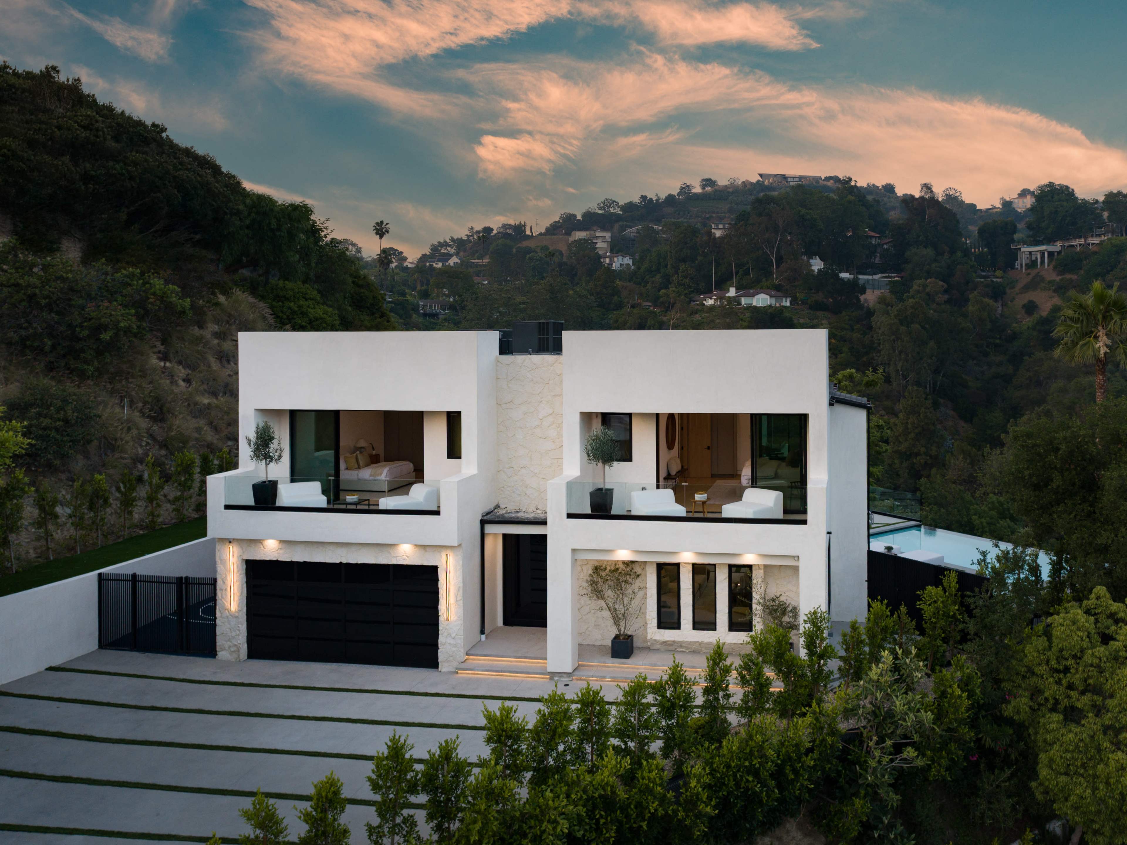 The image shows a modern two-story white house situated on a hillside, featuring large windows, a two-car garage, and landscaped greenery in the foreground.