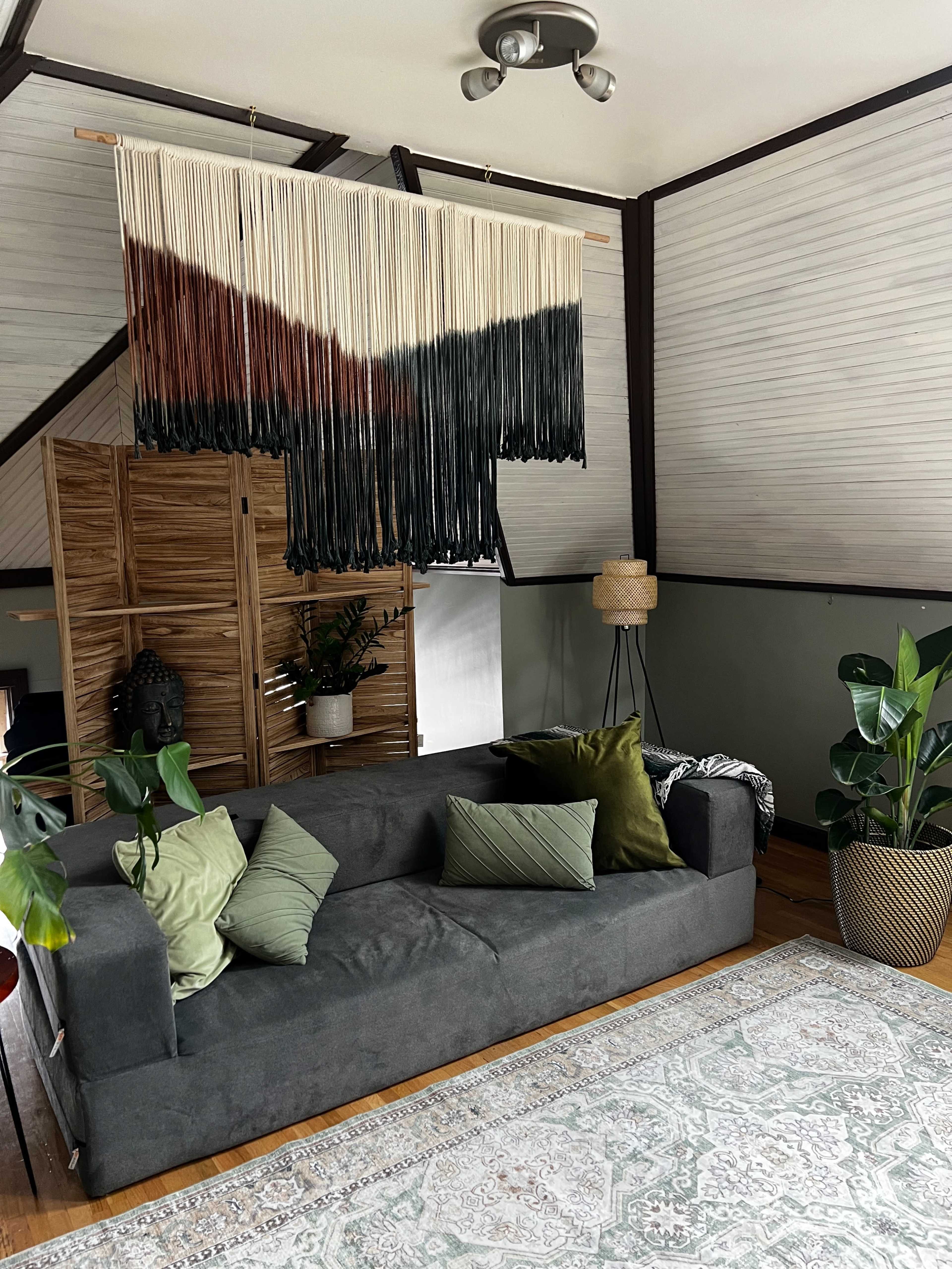 The image shows a living room with a dark green sofa, a large decorative wall hanging, and various plants arranged around a patterned rug.