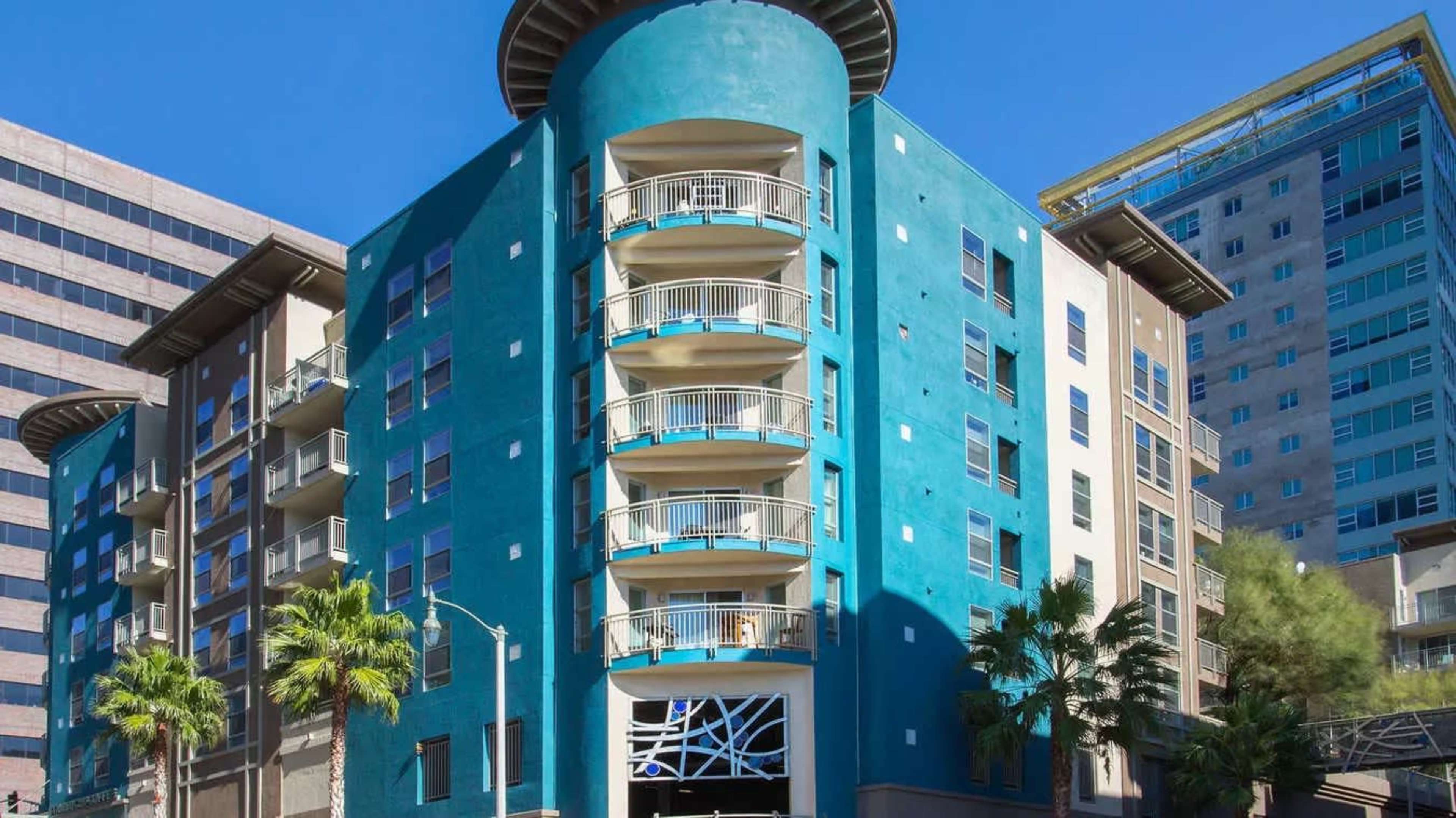 The image shows a modern, multi-story building with a distinctive turquoise exterior, featuring rounded balconies and palm trees in the foreground.