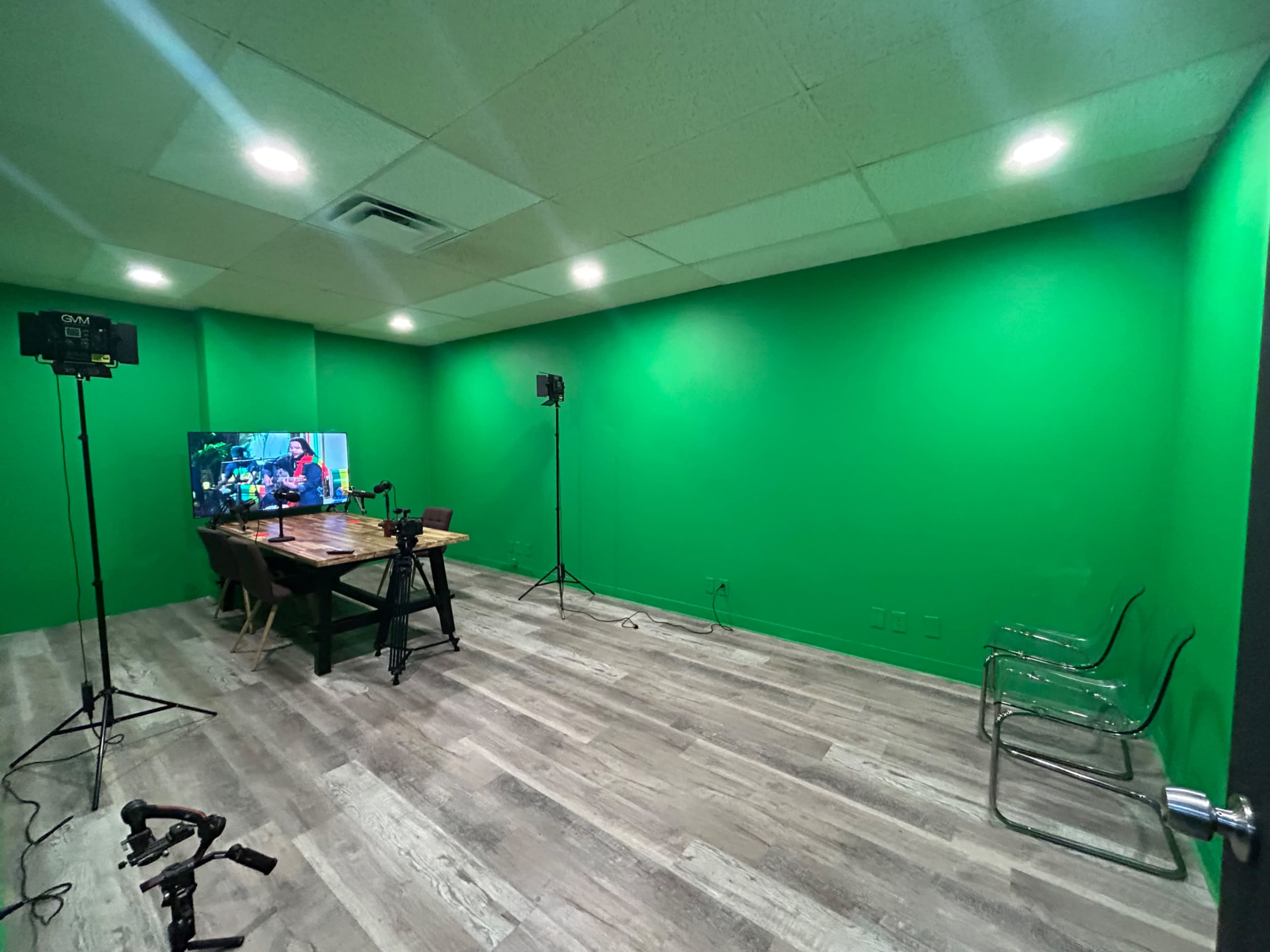 The image shows a green room with a table, transparent chairs, a television displaying a colorful scene, and lighting equipment set up for filming.