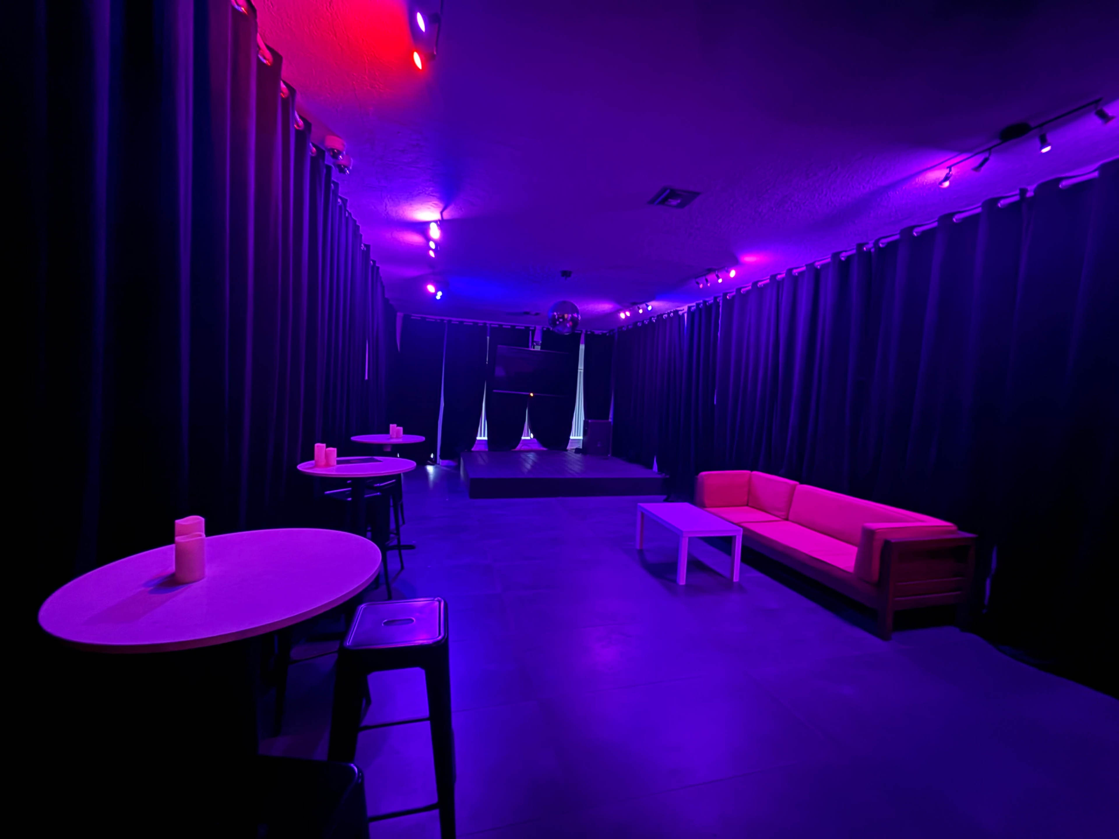 The image shows a dimly lit room with purple lighting, featuring a pink couch, white table, and two small round tables against black curtains.