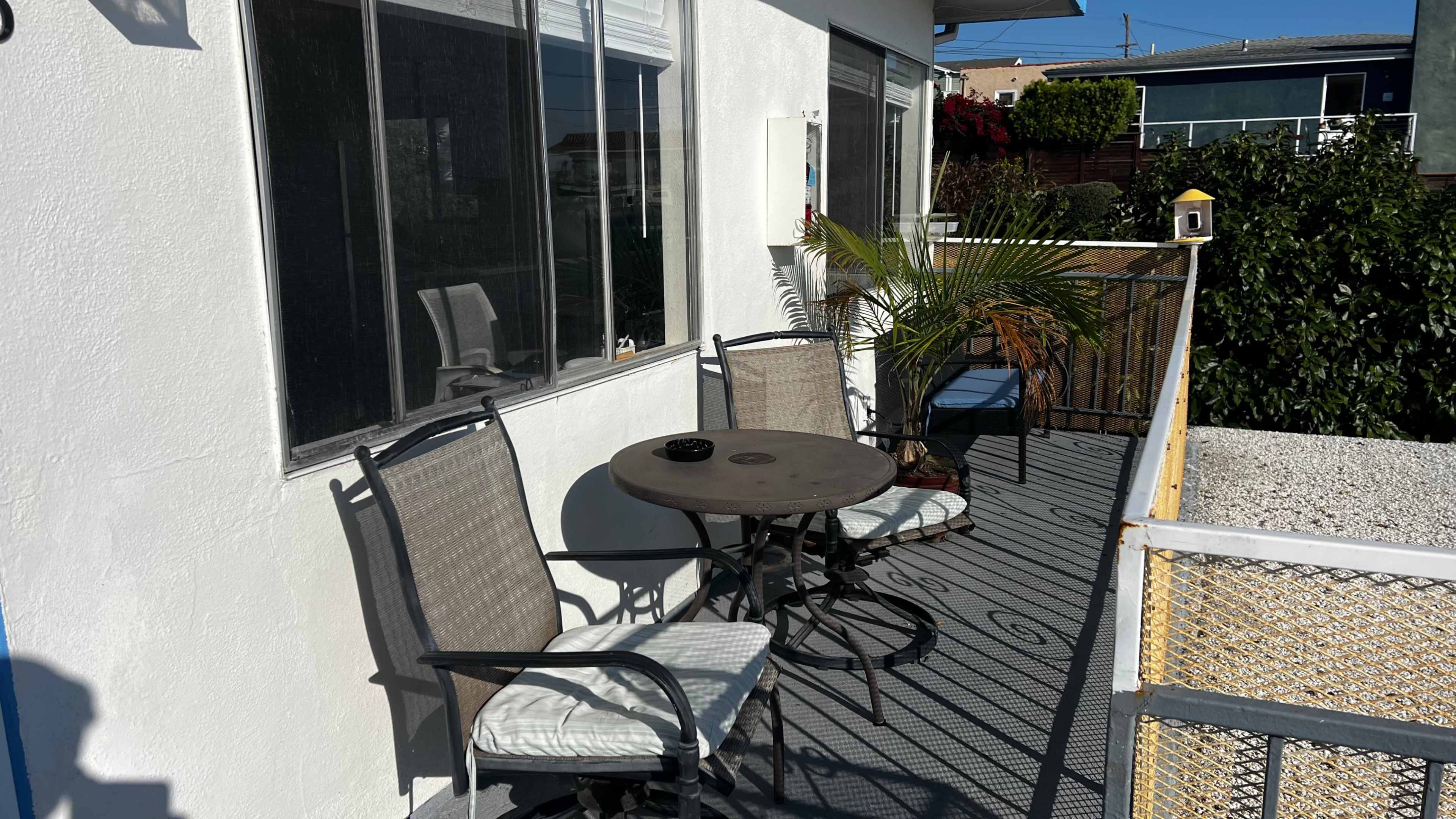 The image shows a small balcony with two chairs and a round table, surrounded by plants and a view of the outdoors.