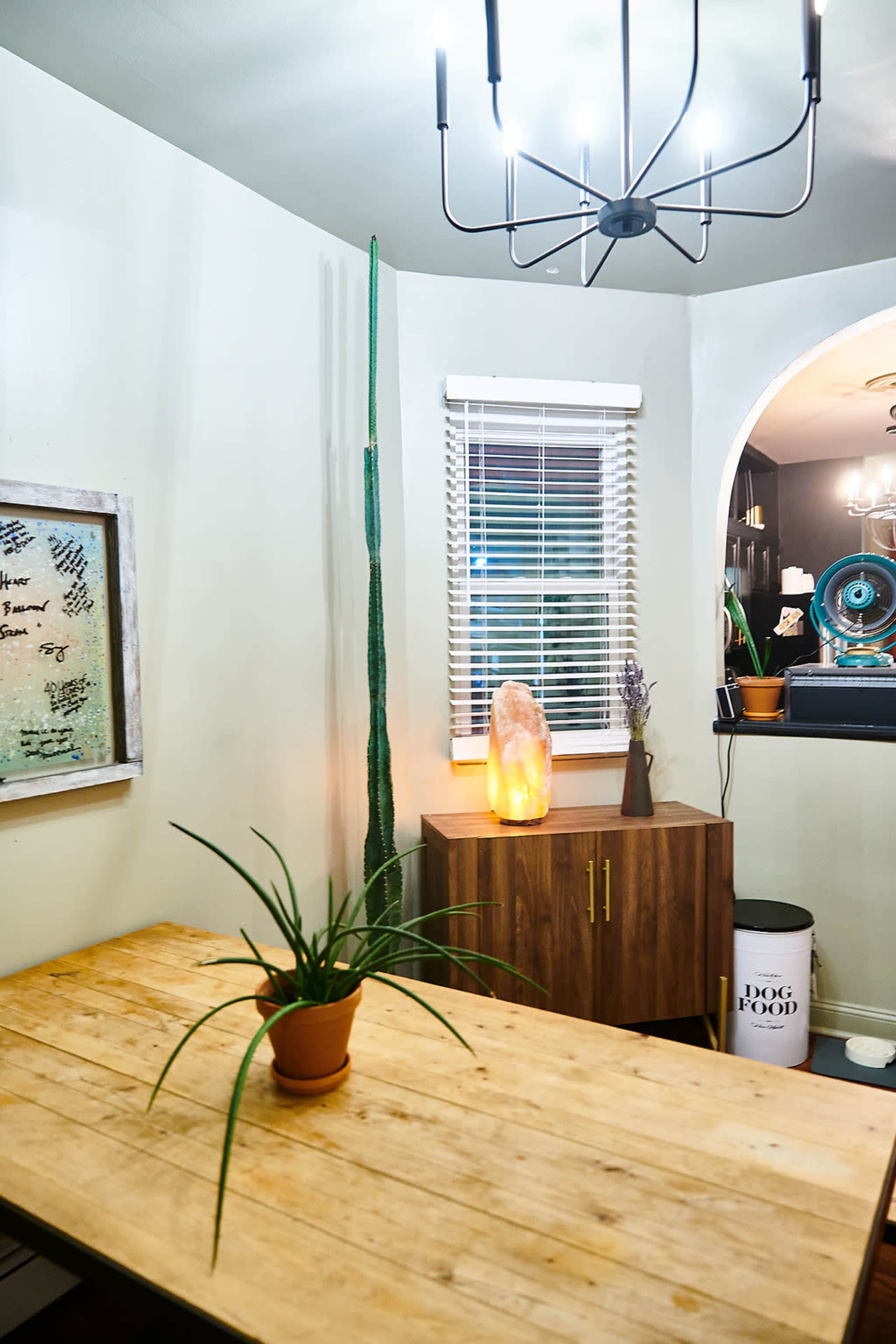 A corner of a dining area features a wooden table with a potted plant, a cabinet, and a Himalayan salt lamp, illuminated by a modern chandelier.