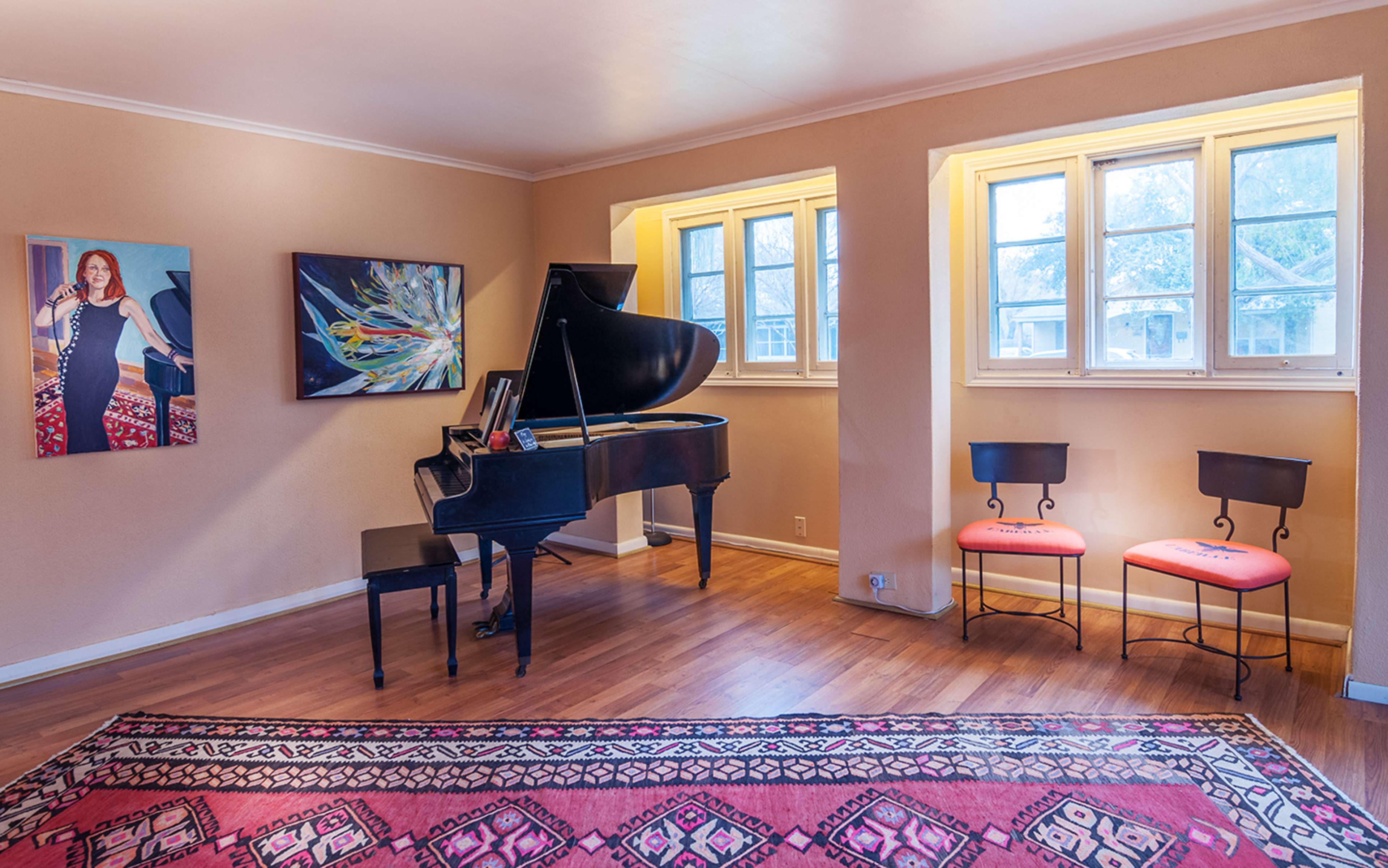 A room featuring a grand piano, two chairs, and framed artwork on the walls, with windows allowing natural light to enter.