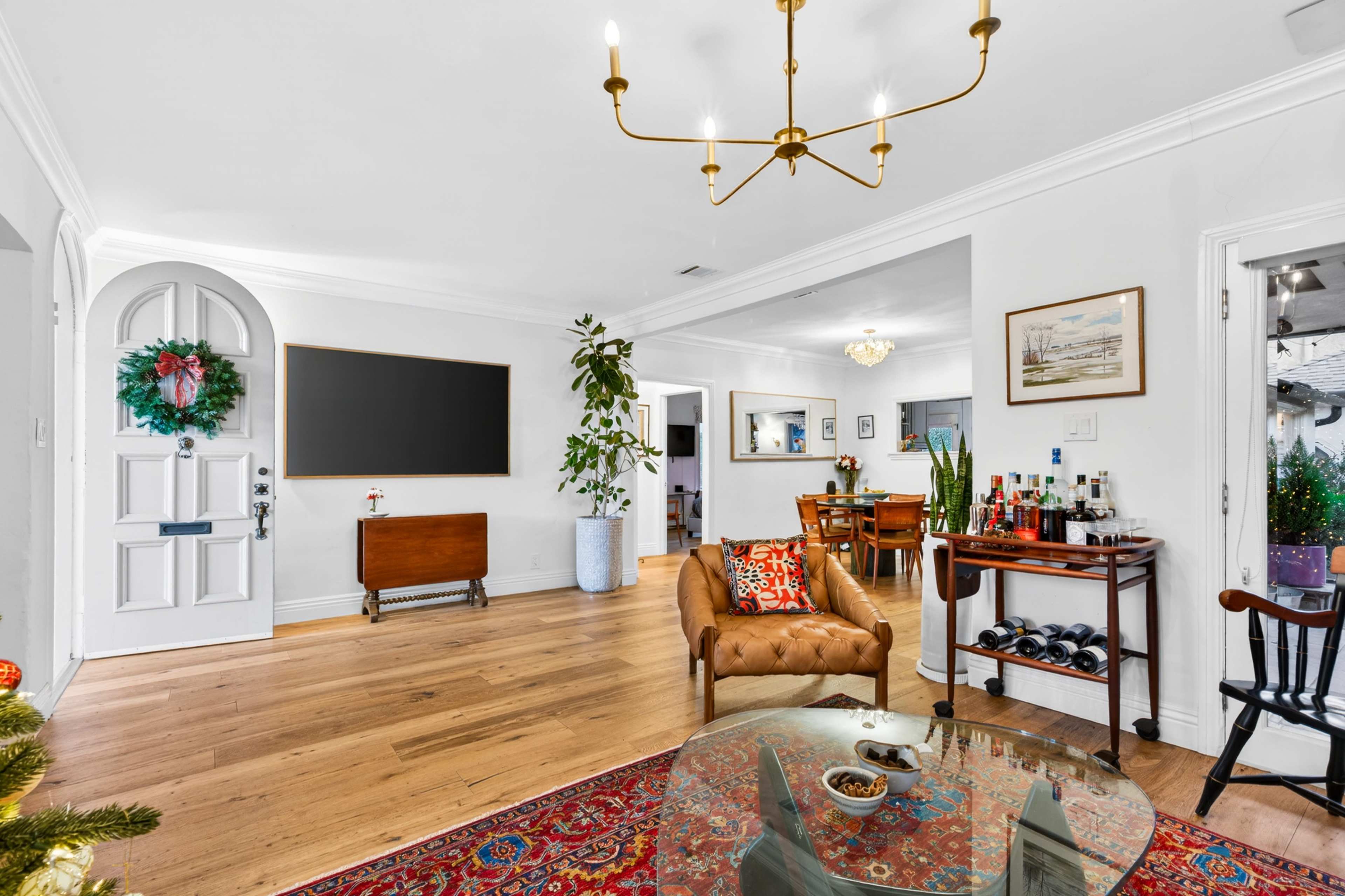 A living room features a cozy seating area with a chesterfield sofa, a large wall-mounted chalkboard, and a dining area in the background, adorned with decorative plants and a variety of furnishings.