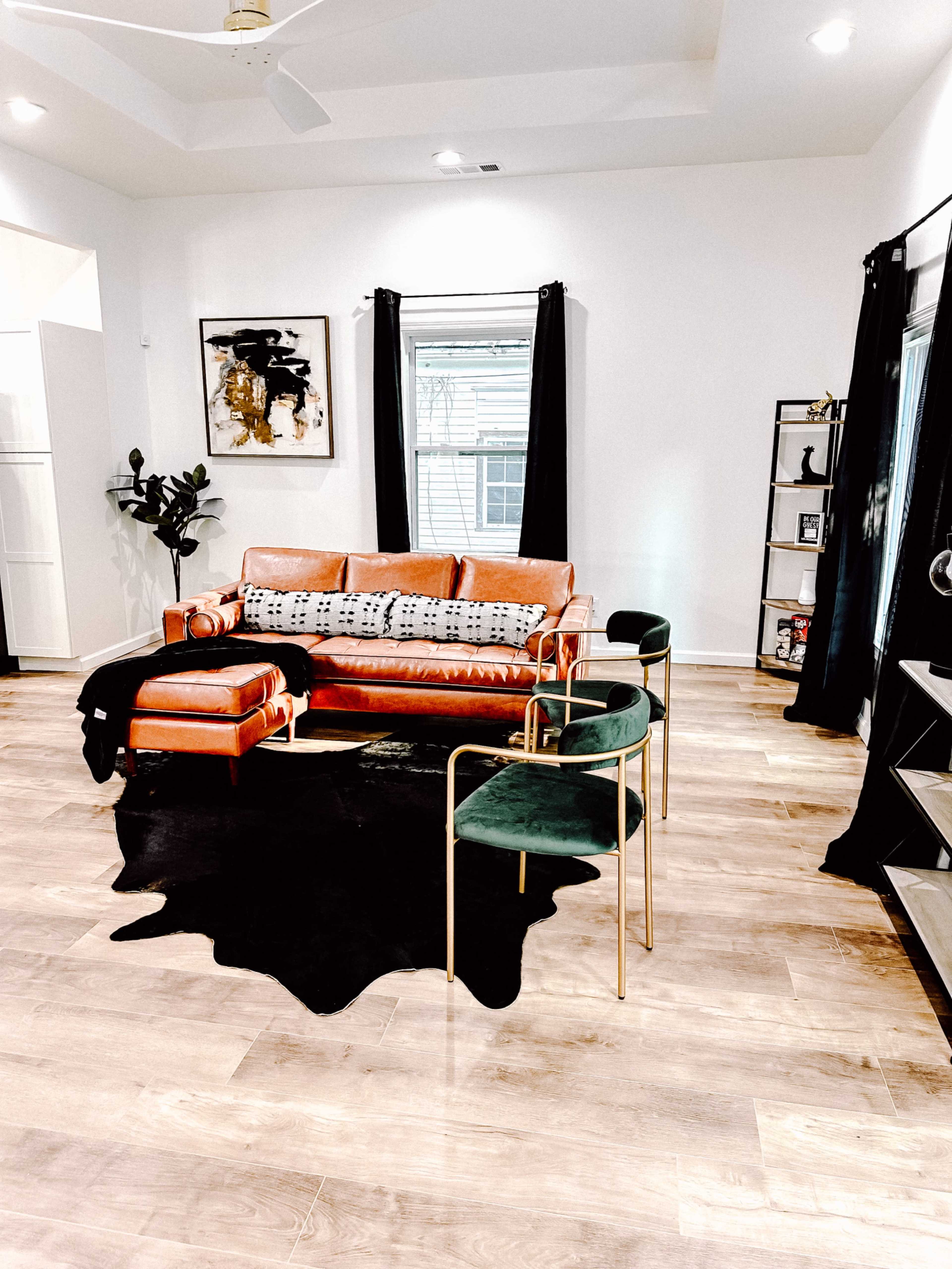 A modern living room with a brown leather sofa, two green chairs, a black rug, and large windows draped with black curtains.