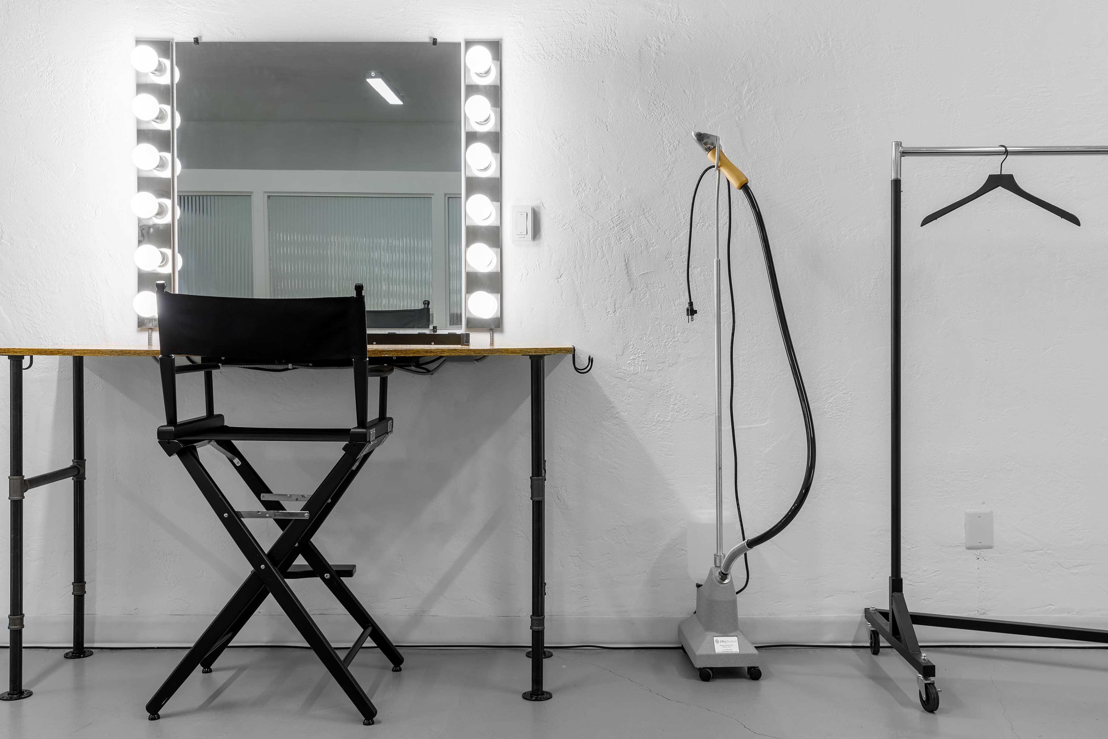 A makeup station with a lighted mirror, a director's chair, a garment rack, and a steamer is set up in a minimalist room.