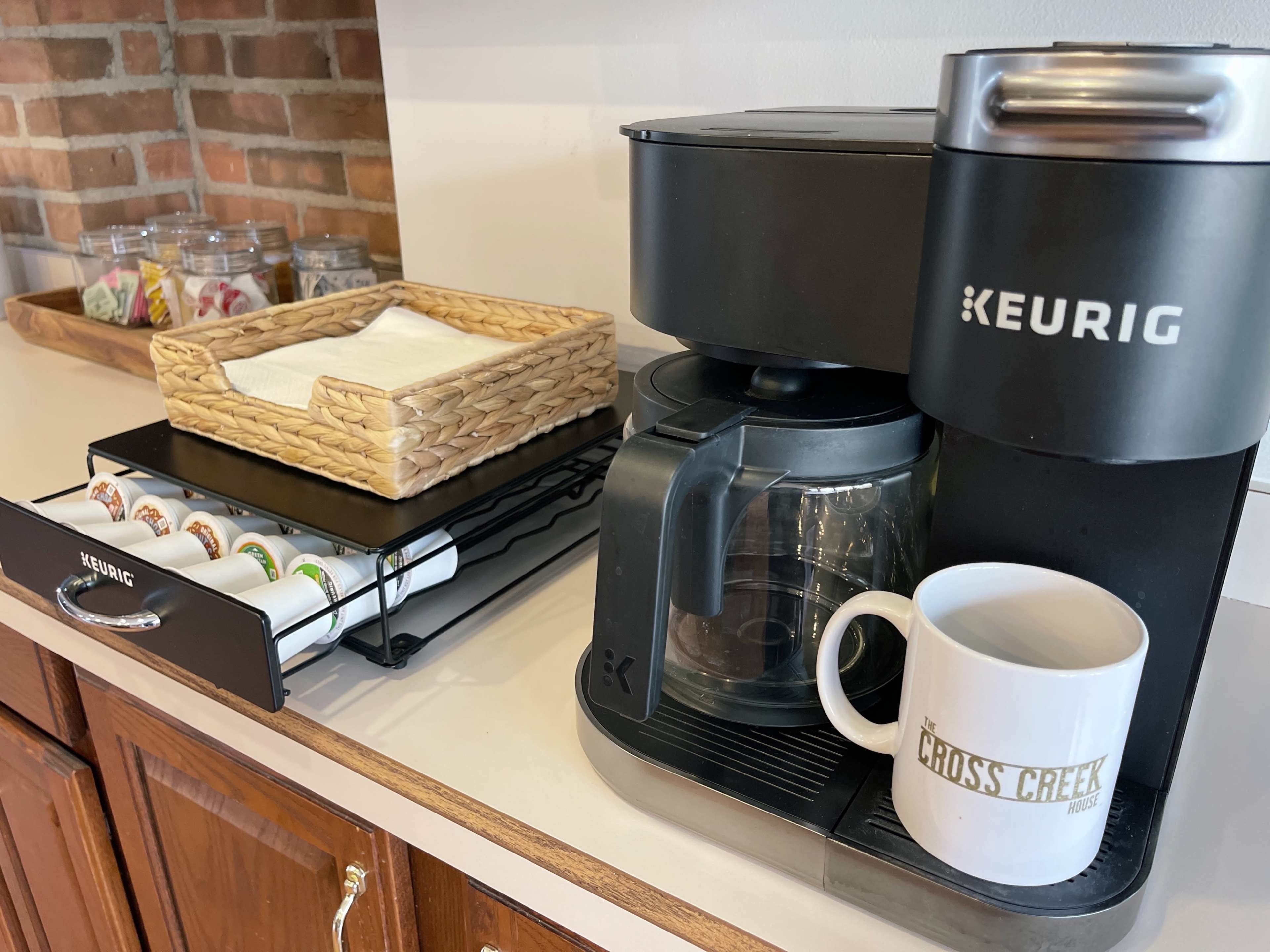 A Keurig coffee maker sits on a countertop next to a basket of coffee pods and a white mug with the text "CROSS CREEK."
