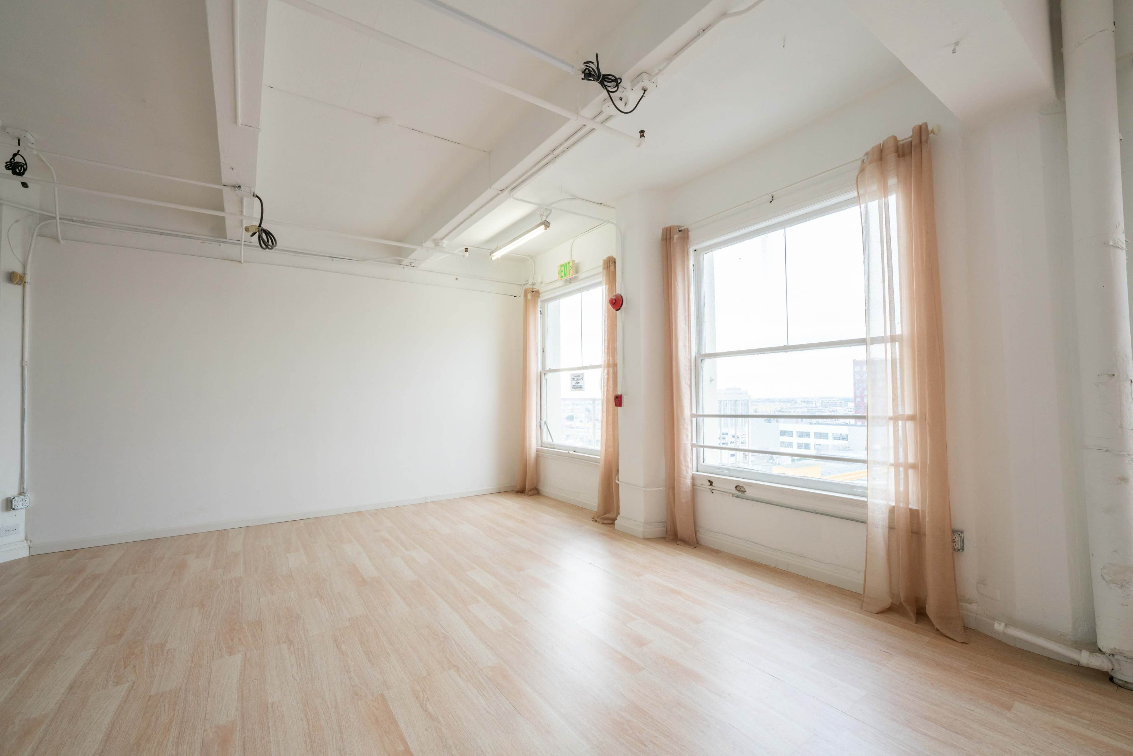 A spacious, empty room with hardwood flooring, large windows, and sheer curtains.