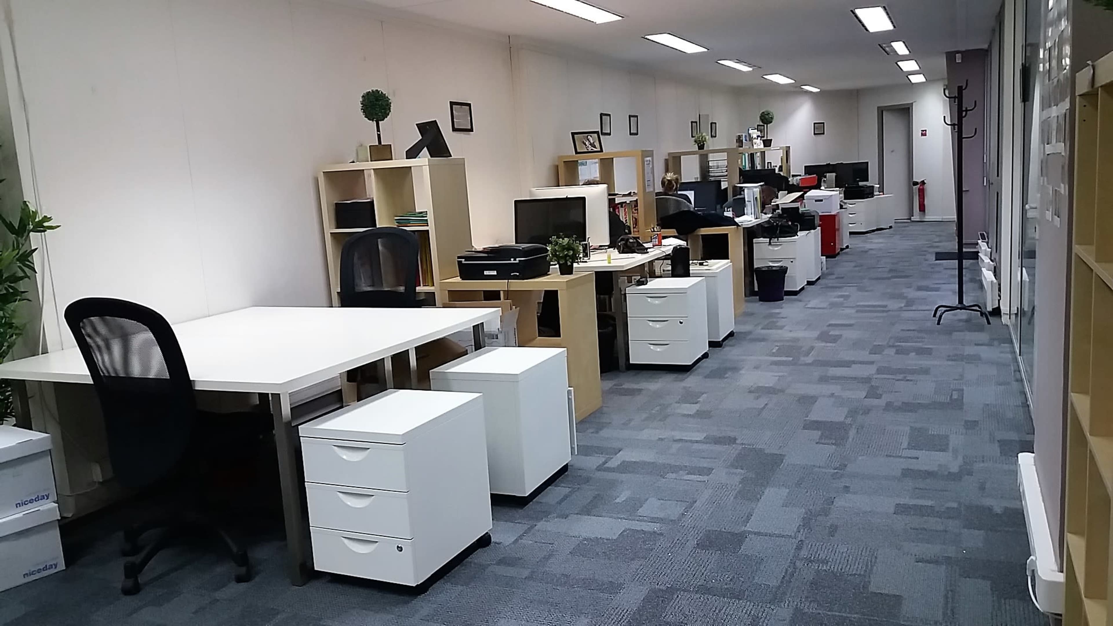 A modern office space with desks, chairs, computer workstations, and storage units arranged along a long corridor.