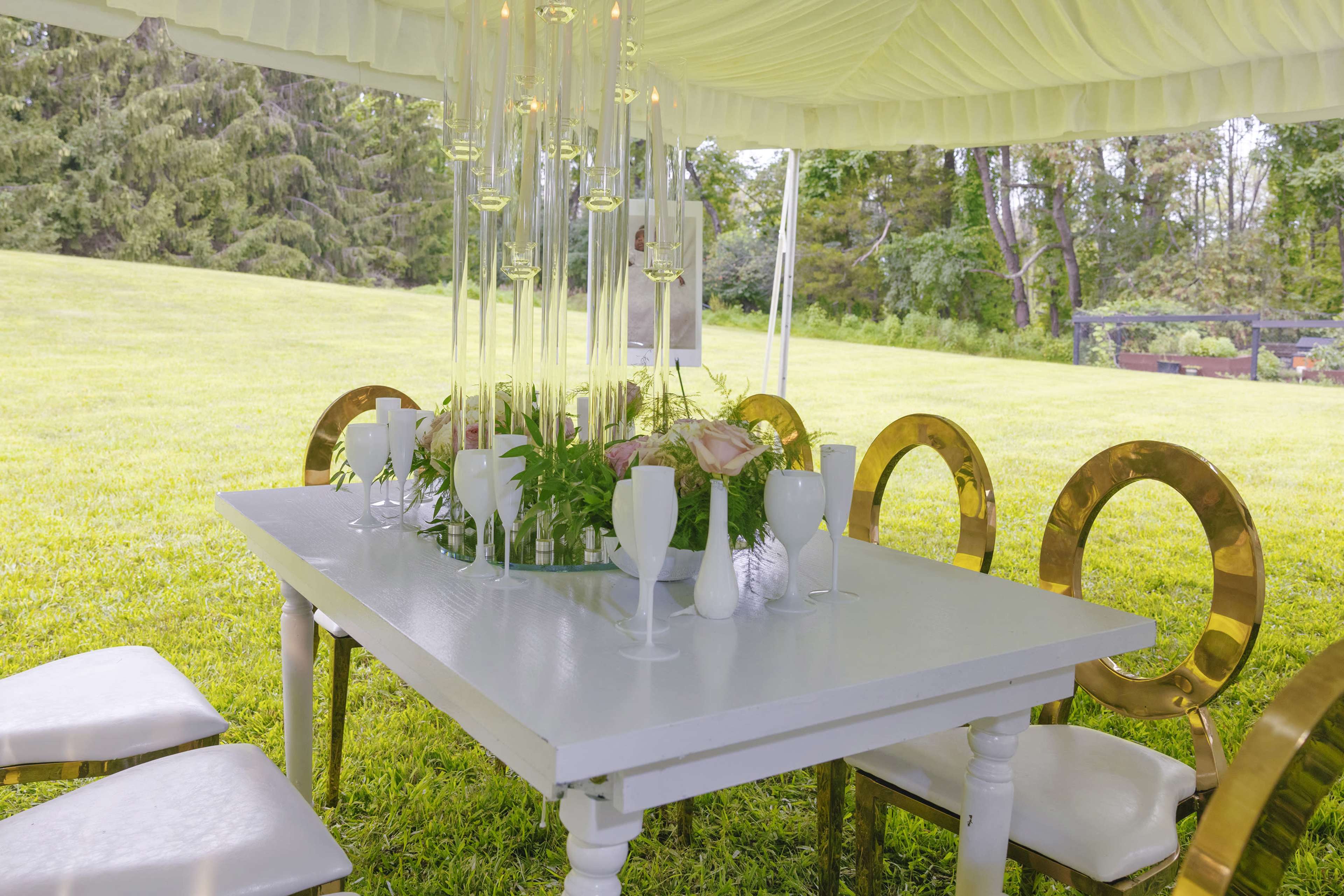 A white table is set under a tent with gold-accented chairs and a modern floral centerpiece amidst a green lawn.