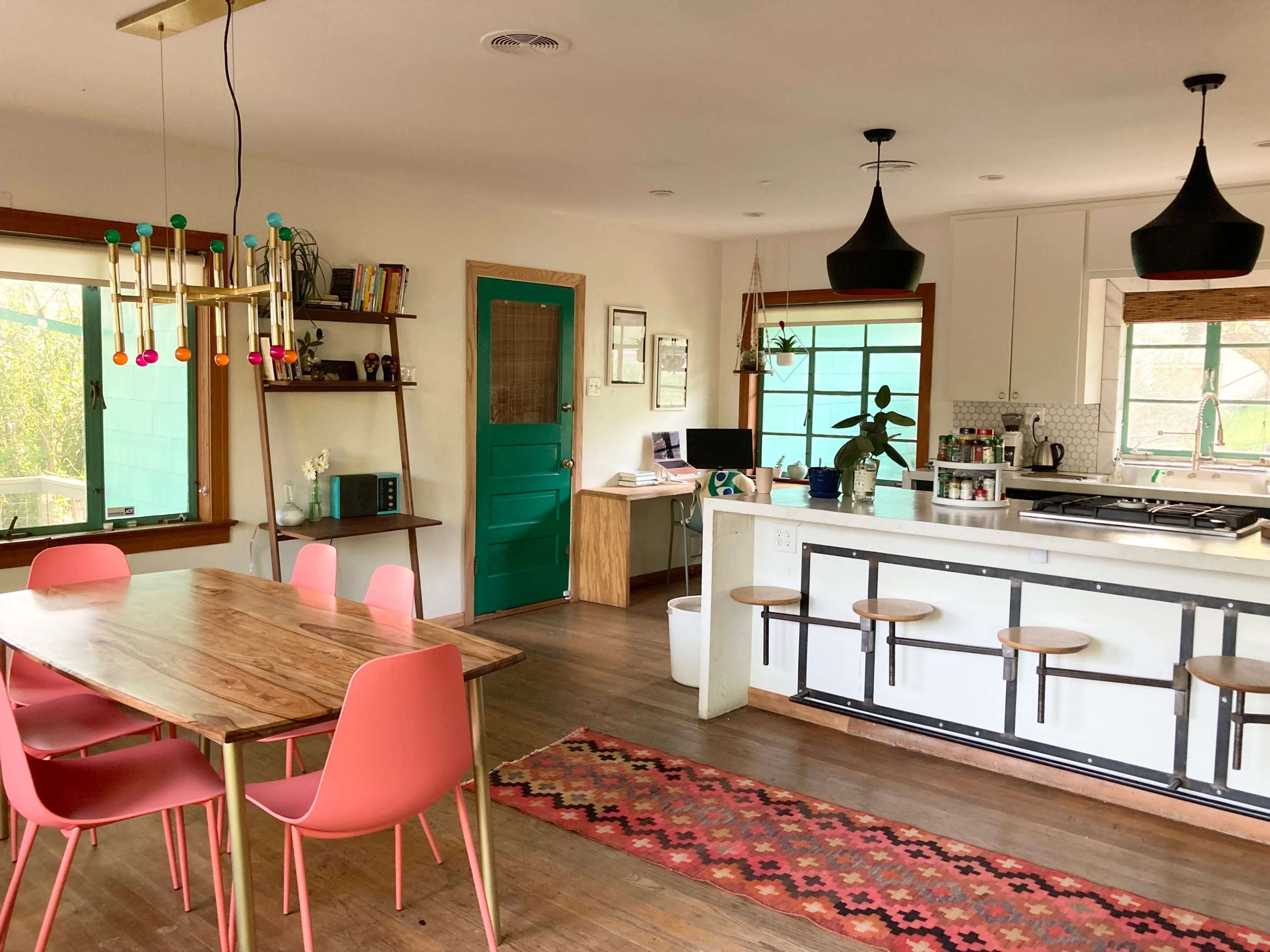 A modern kitchen and dining area features a wooden table with colorful chairs, a green door, and a long kitchen island with stools.