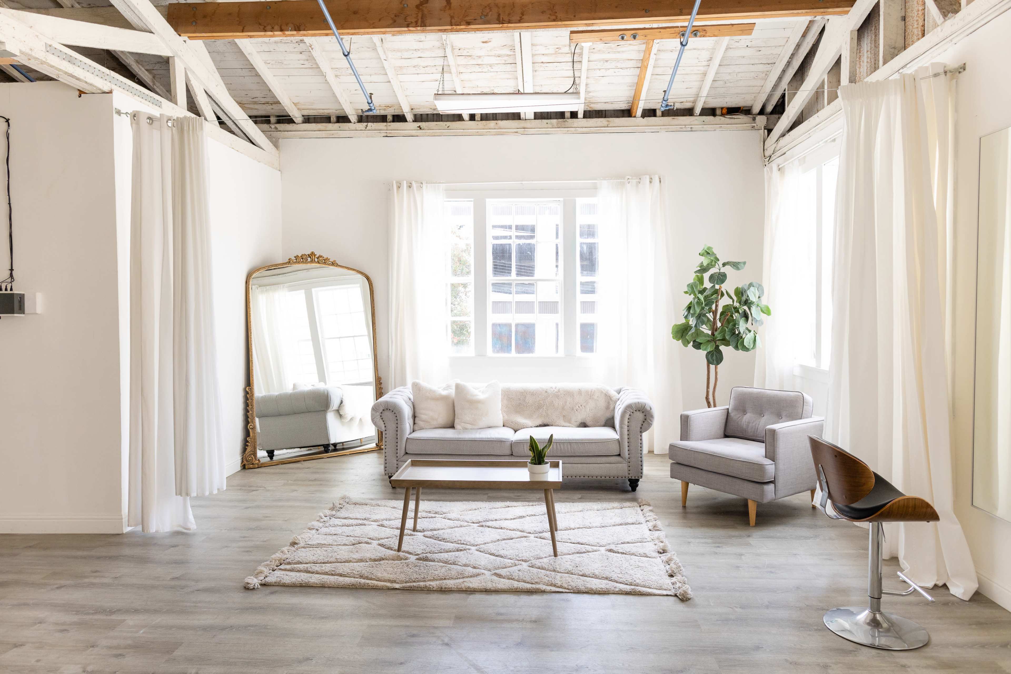 A spacious, well-lit living room featuring a sofa, an armchair, a coffee table, and a large mirror against a backdrop of white walls and wooden beams.