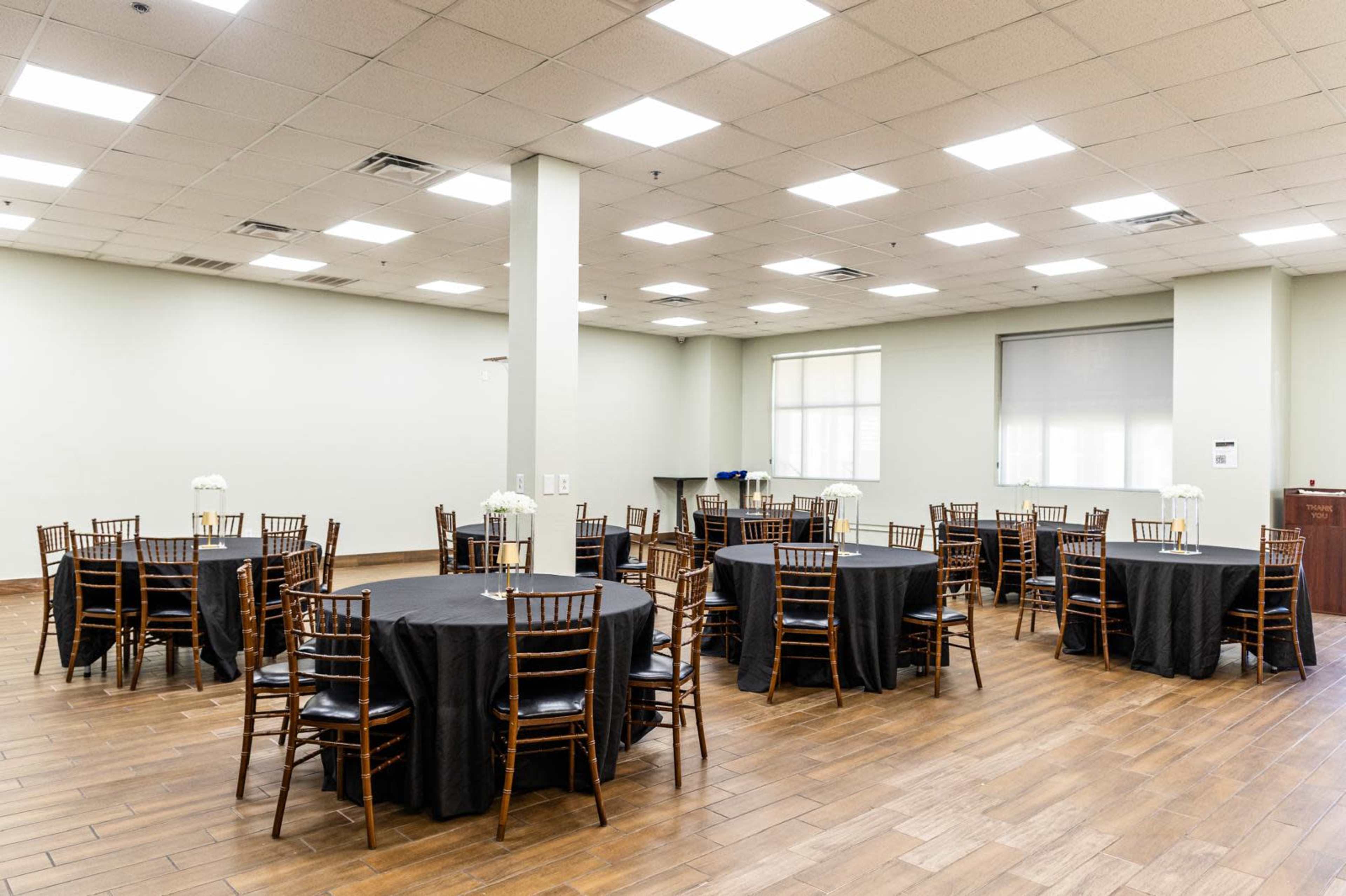 The image shows a spacious event room with several round tables covered in black tablecloths and wooden chairs arranged around them, under bright overhead lights.
