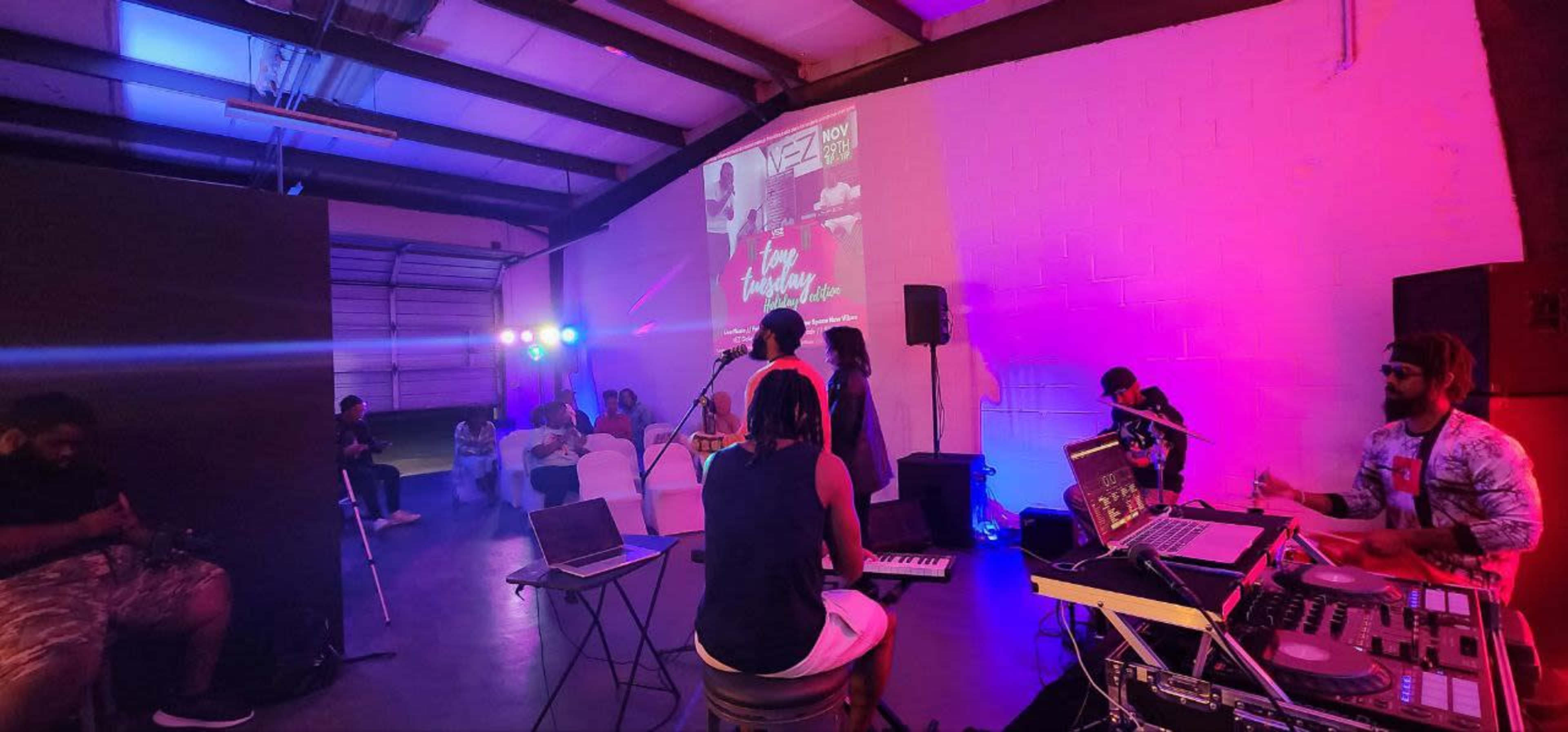 A group of musicians perform in a brightly lit warehouse space with a DJ setup and audience members seated nearby.