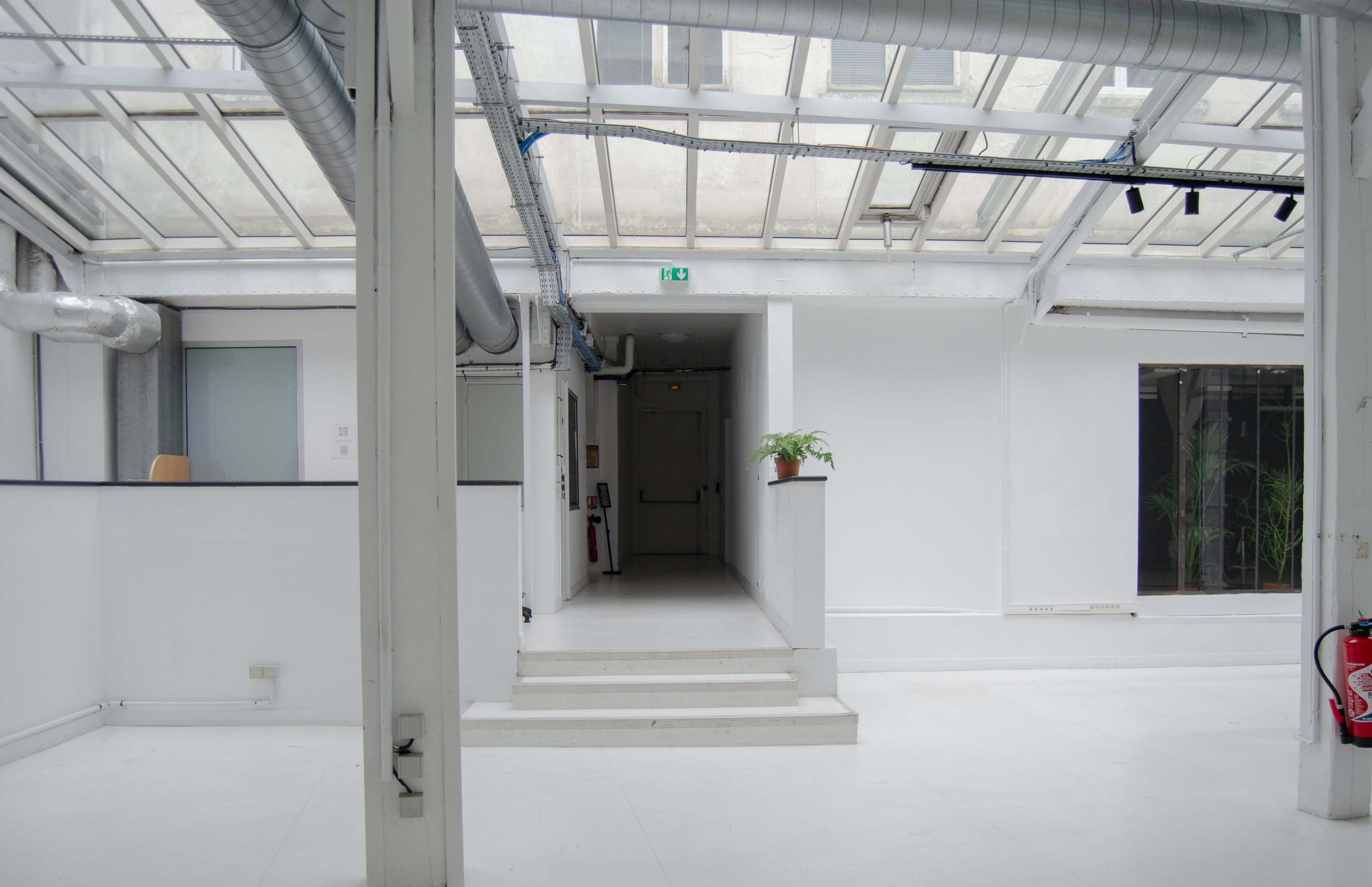 The image shows a minimalist interior space with white walls, a staircase leading to a higher level, and large windows allowing natural light to enter.