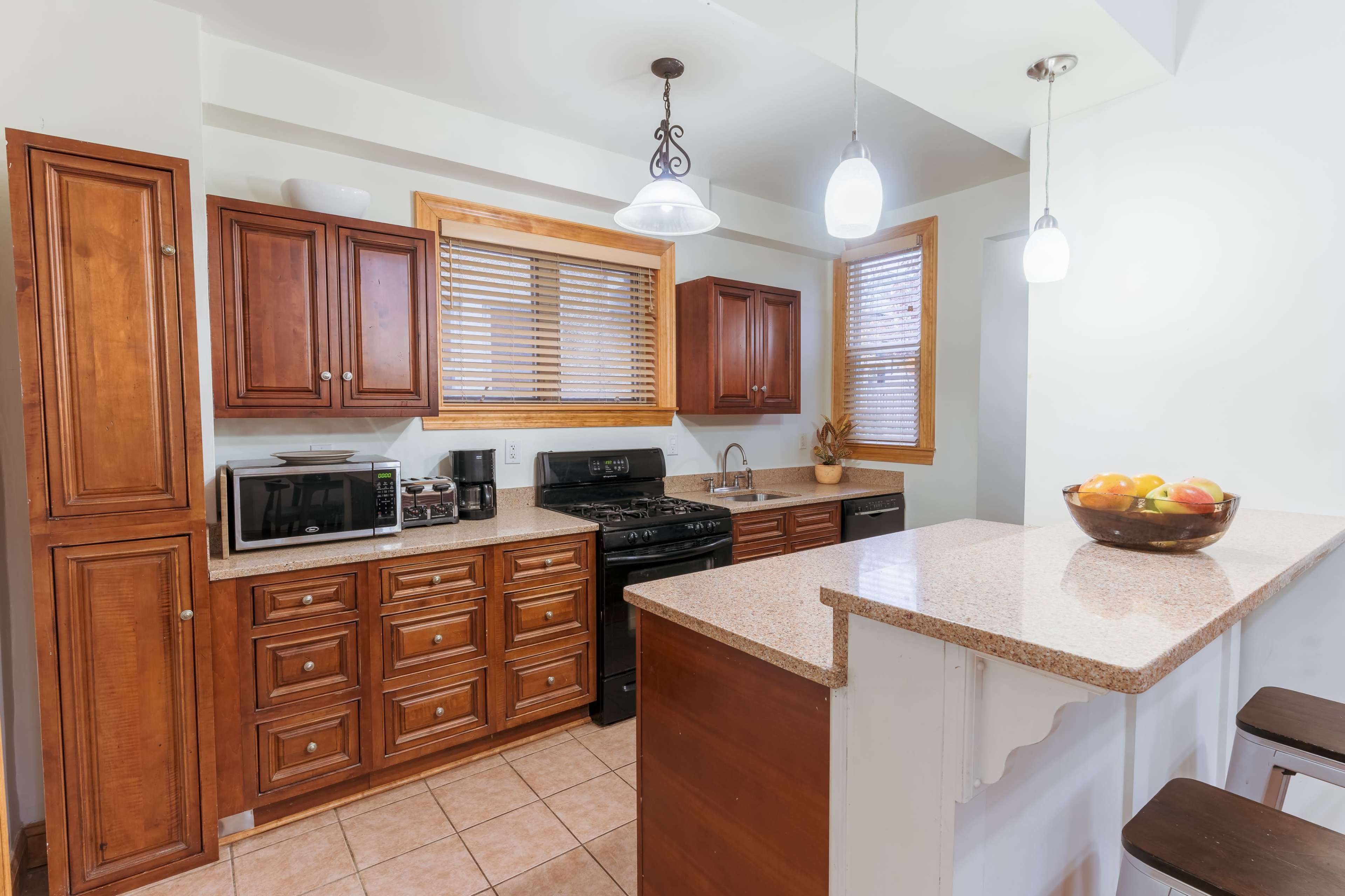 The image shows a kitchen with wooden cabinets, a black stove, a microwave, and a bar area featuring a bowl of fruit.