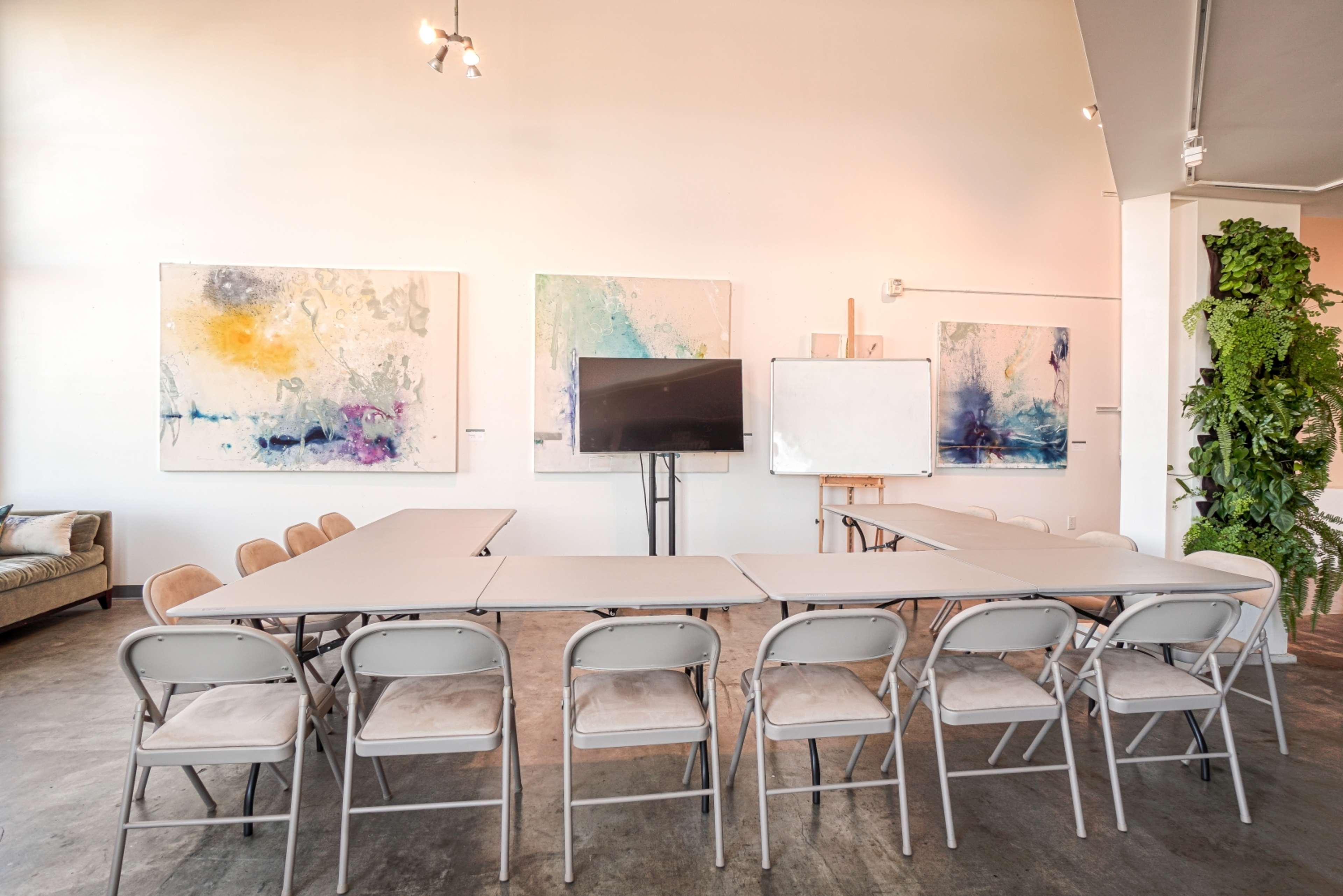A long table with folding chairs is set up in a brightly lit room adorned with large abstract paintings and equipped with a television and a whiteboard.