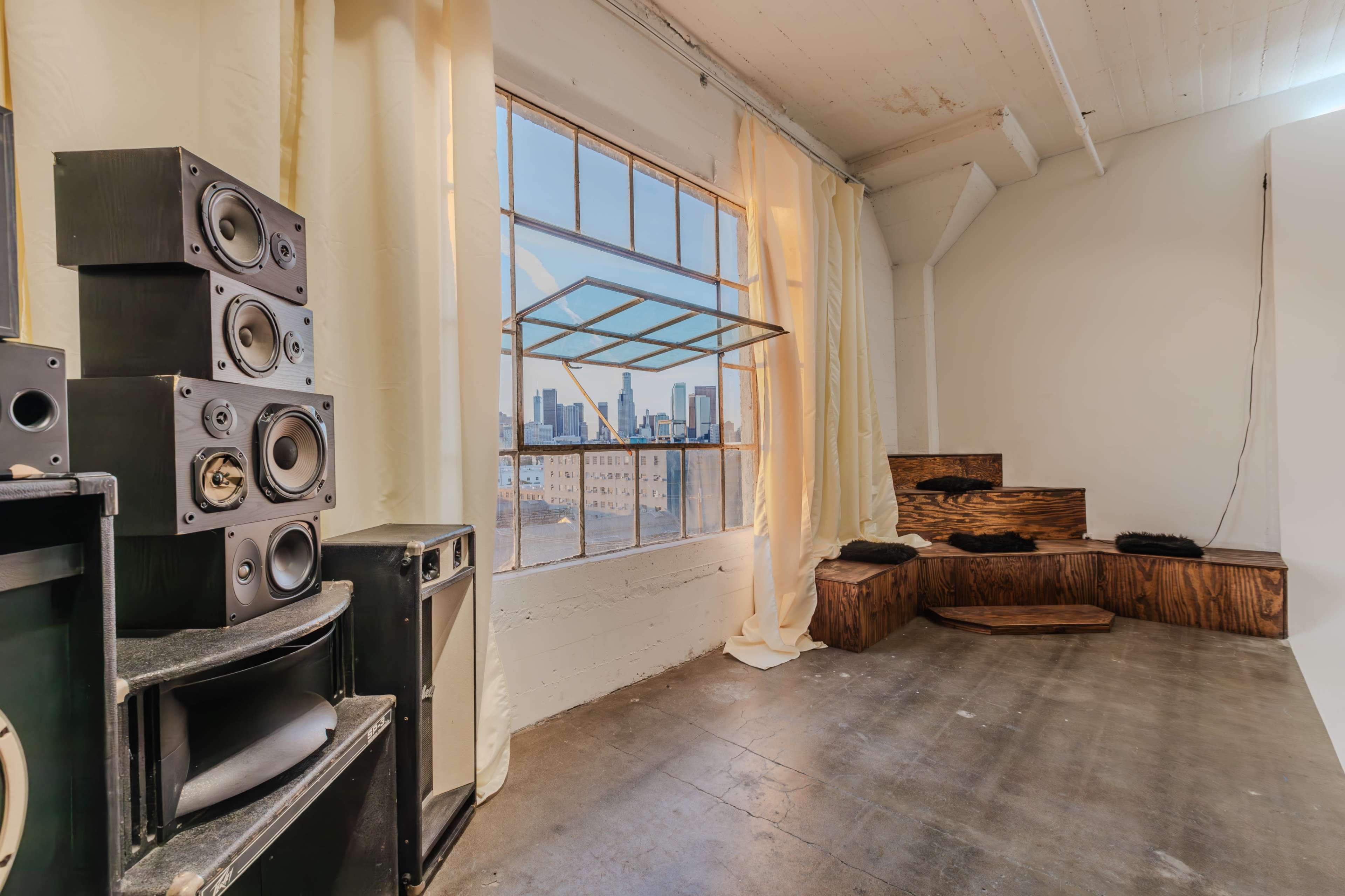 A room with large windows offers a view of a city skyline, and features stacked speakers and wooden seating along the walls.