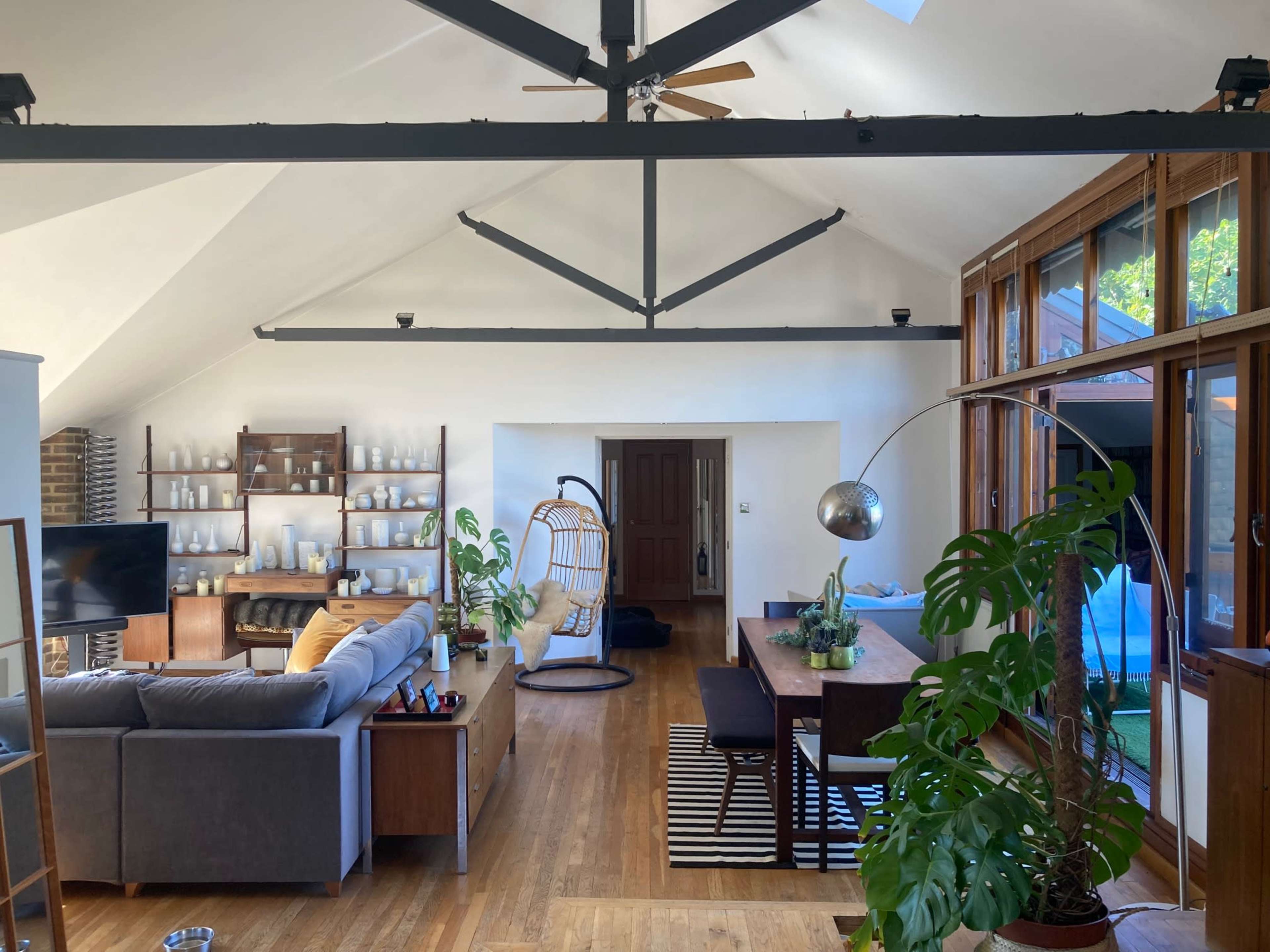 The image shows a spacious, modern living area featuring a gray sofa, dining table, and a hanging chair, with large windows allowing natural light to fill the space.