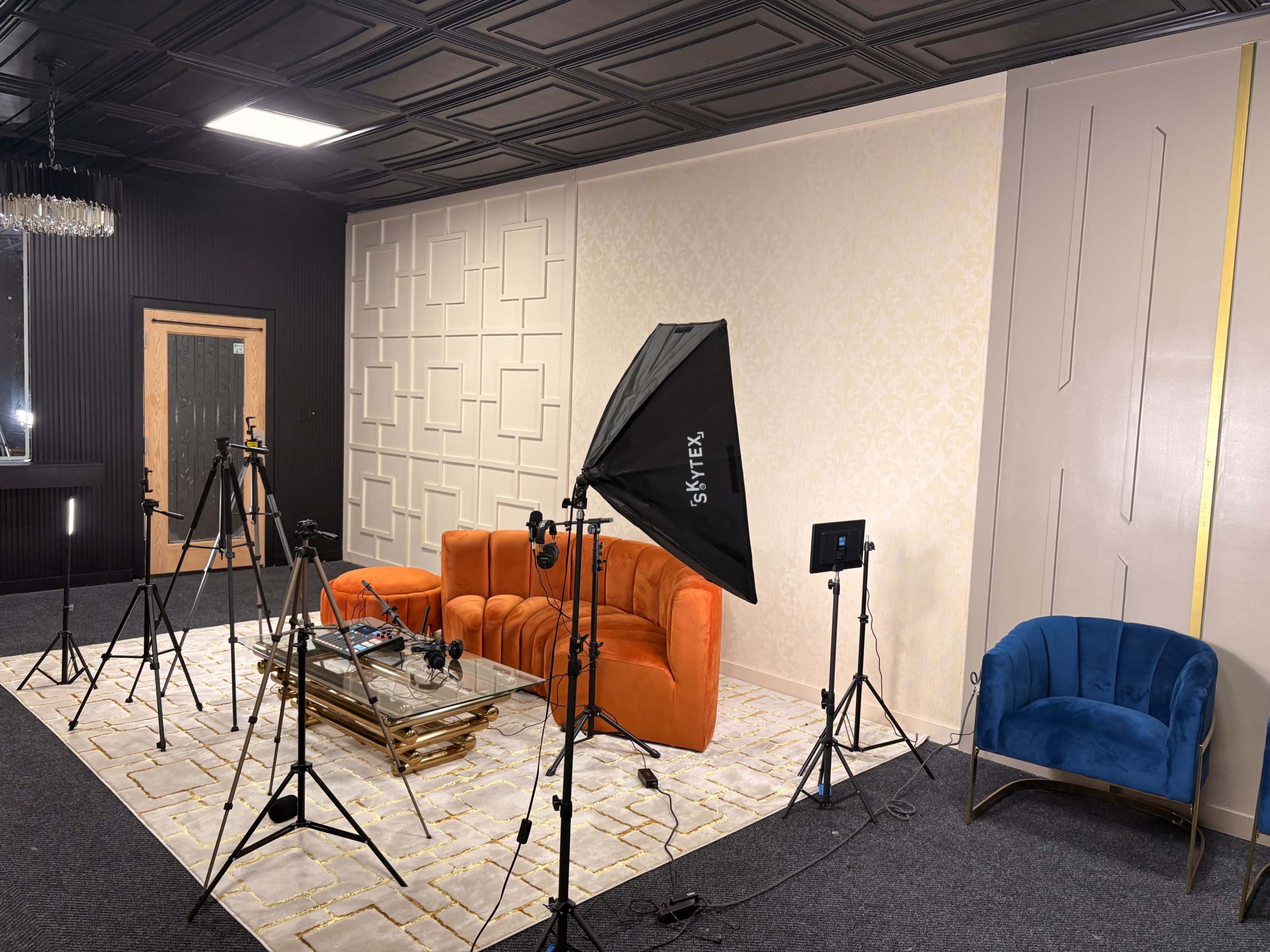 The image shows a studio setup featuring an orange sofa, a blue chair, and multiple cameras and lighting equipment on a patterned rug.