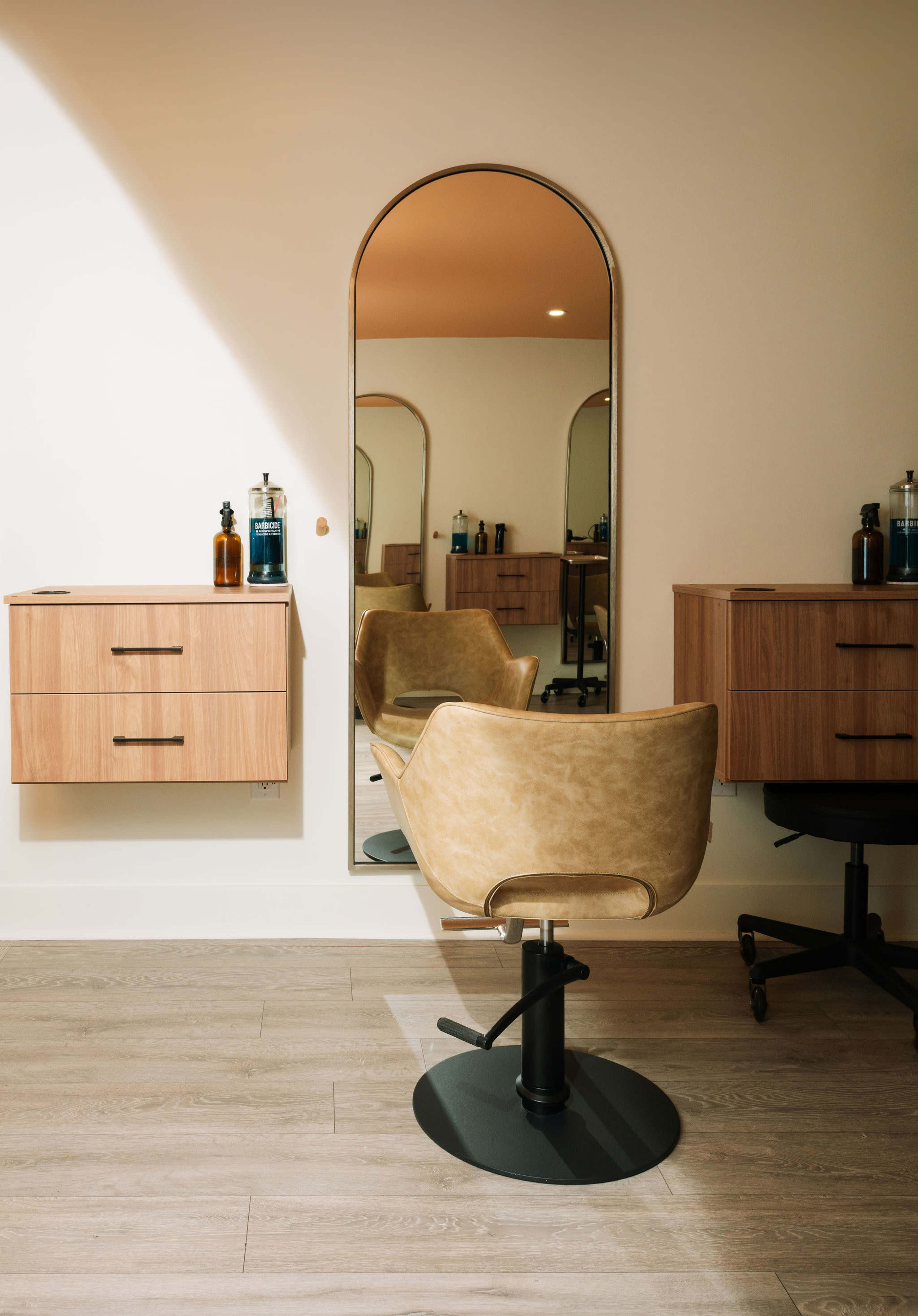 The image shows a modern hair salon interior featuring a stylist chair, two mirrors, and wooden storage cabinets against a light-colored wall.