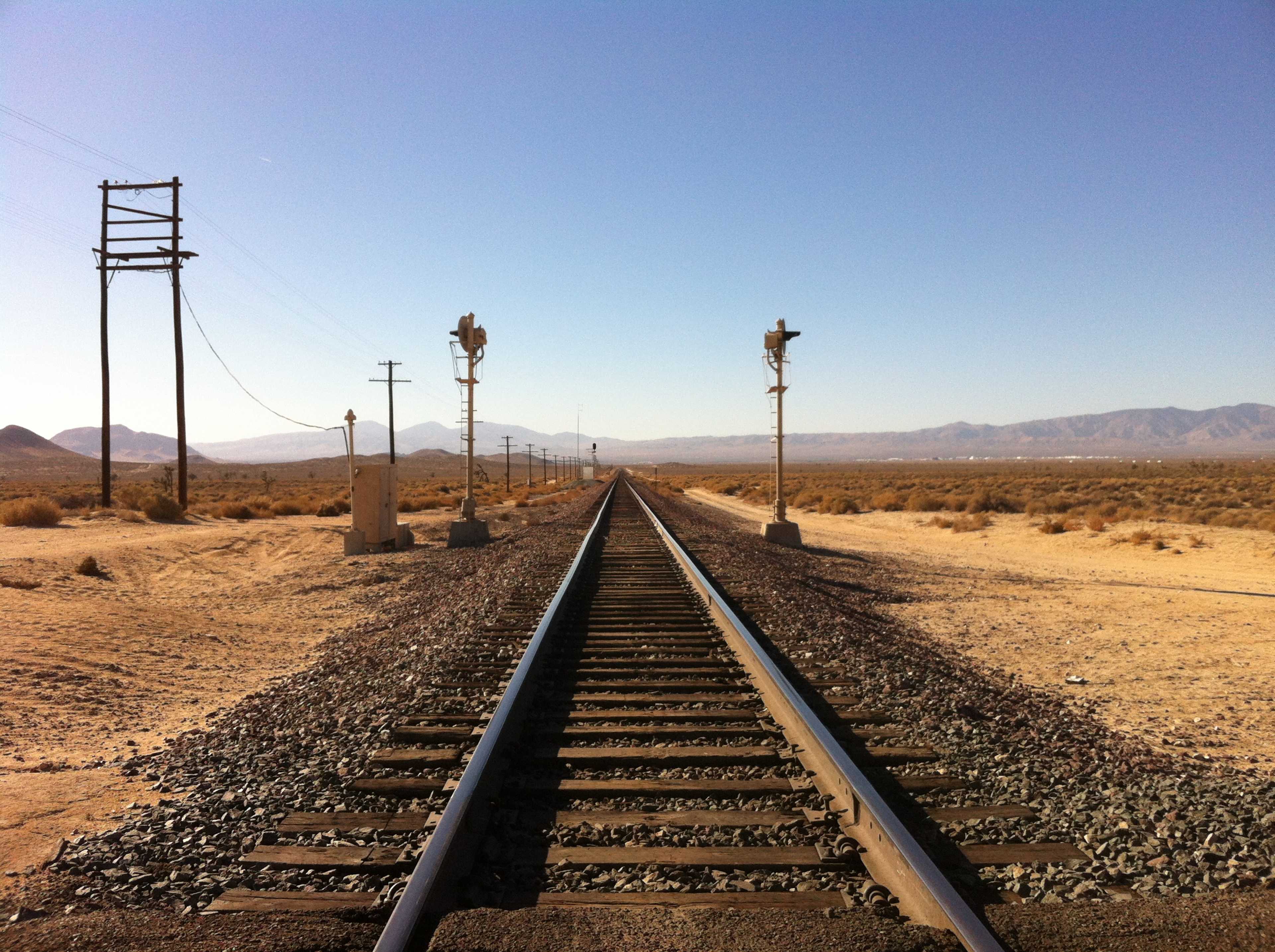 A straight railway track stretching through a desert landscape with telegraph poles and signal lights on either side.