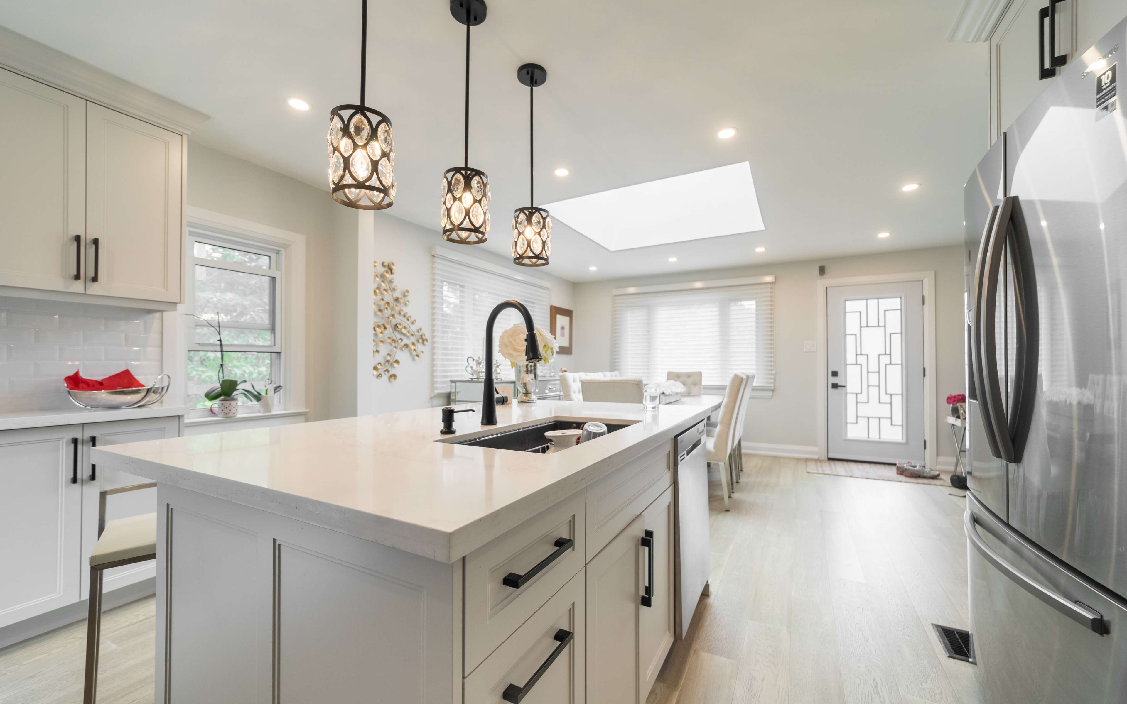 A modern kitchen features a central island with a sink, pendant lighting, and large windows allowing natural light.