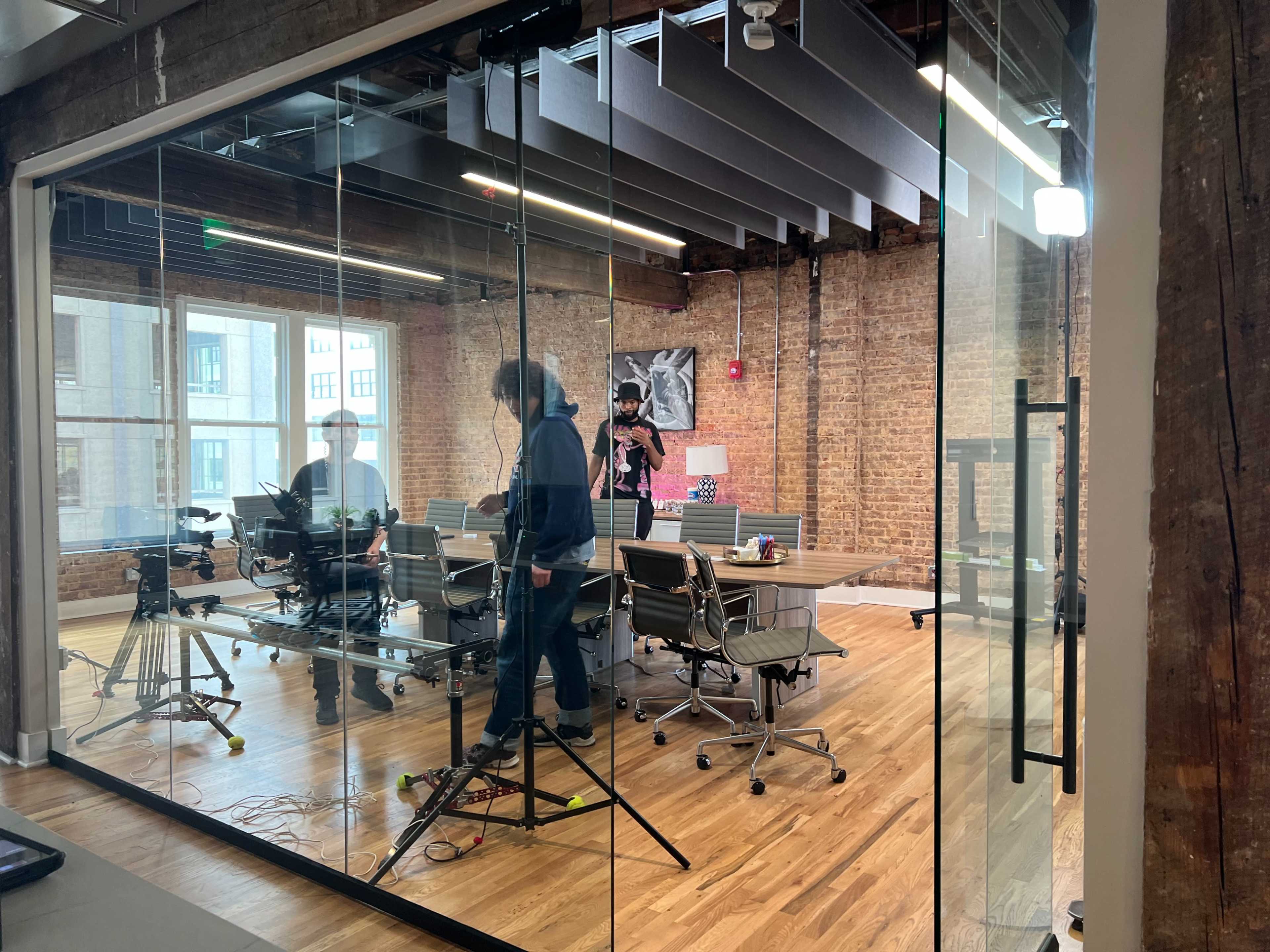 A modern conference room with glass walls, wooden floors, and a long table, where people are setting up equipment for a presentation.