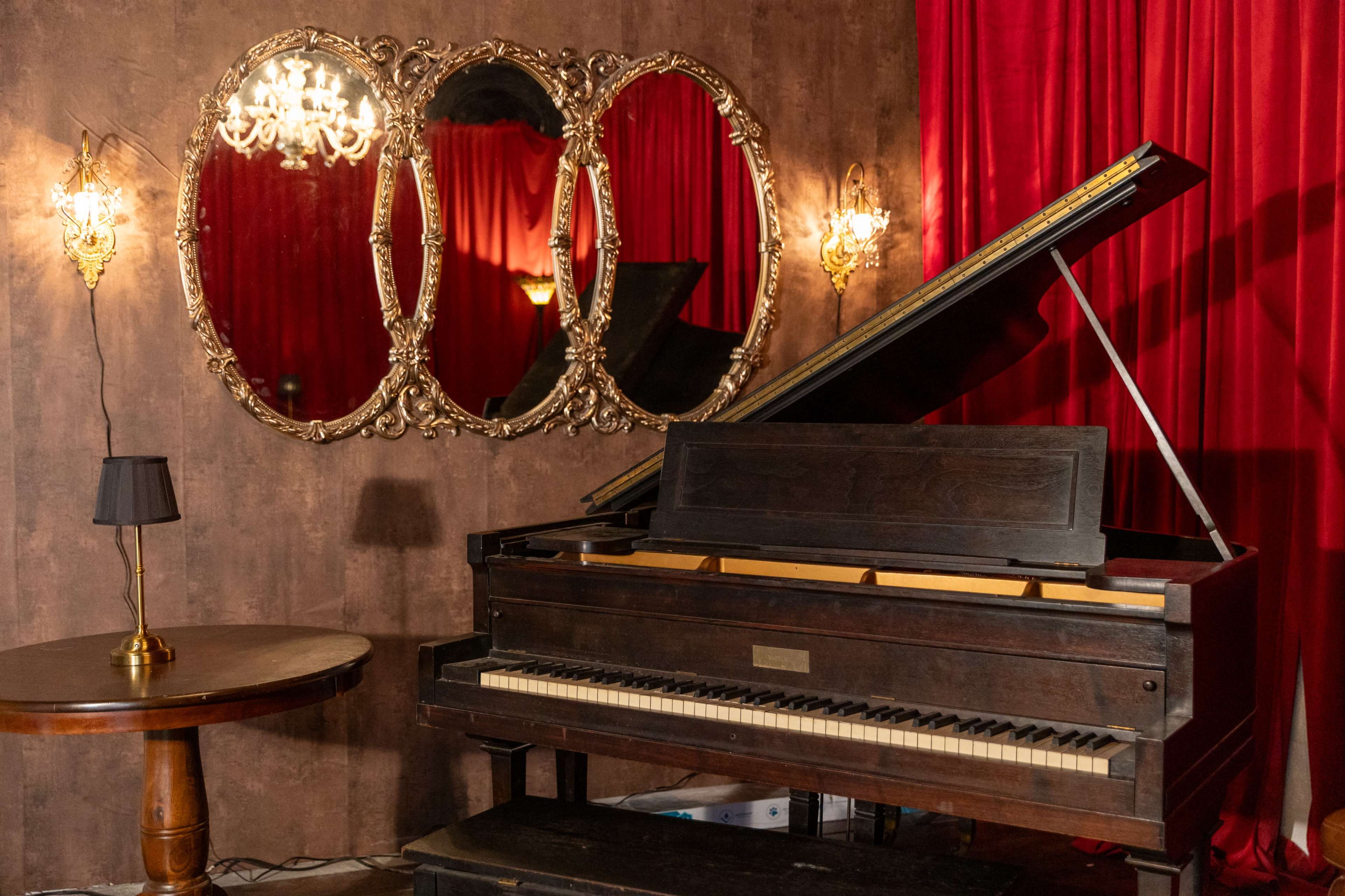 The image shows a vintage piano beside a round wooden table, with ornate mirrors reflecting a red velvet backdrop and decorative lighting.