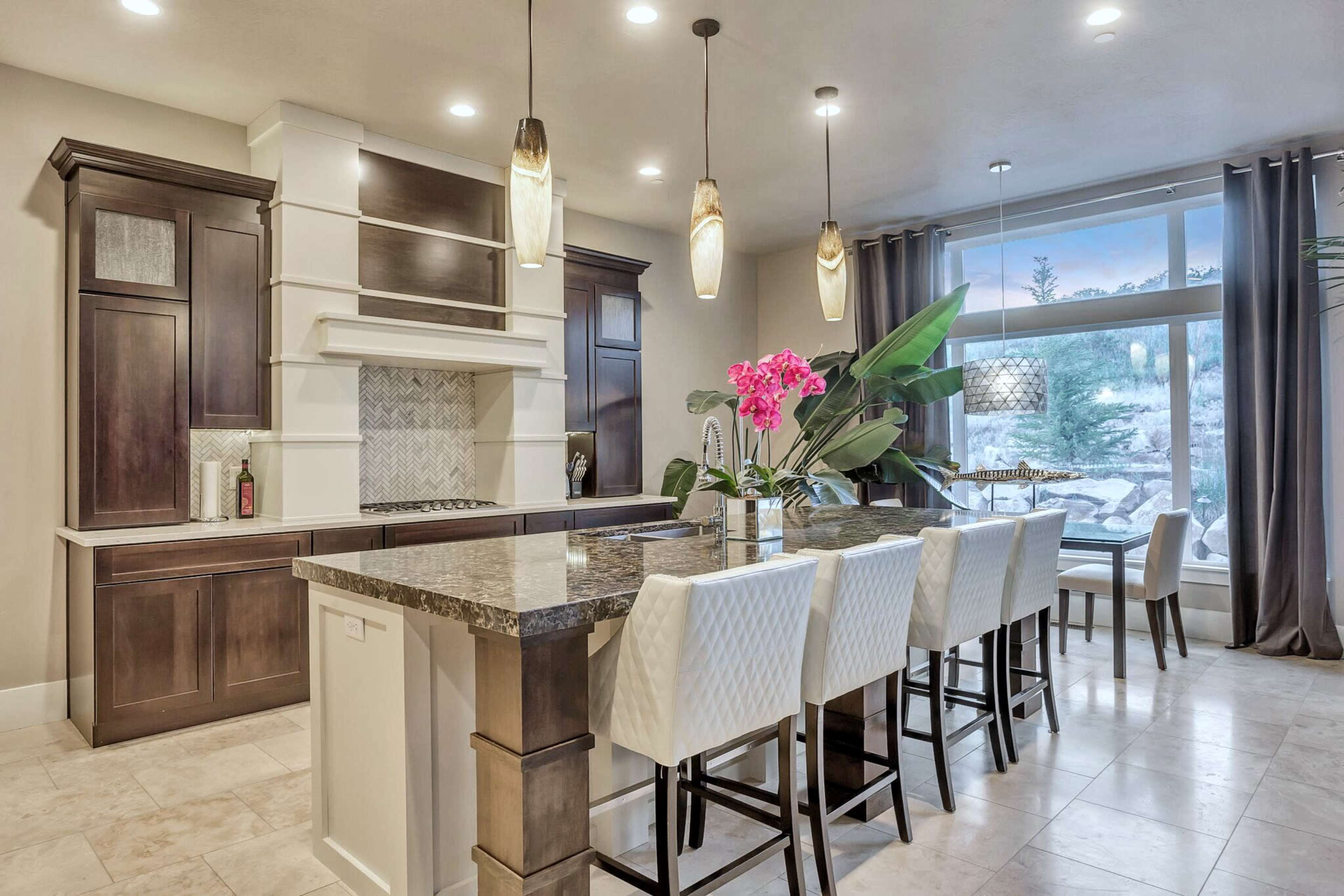 A modern kitchen features dark wood cabinetry, a large island with a granite countertop, and bar seating with white chairs, accented by pink orchids and large windows.