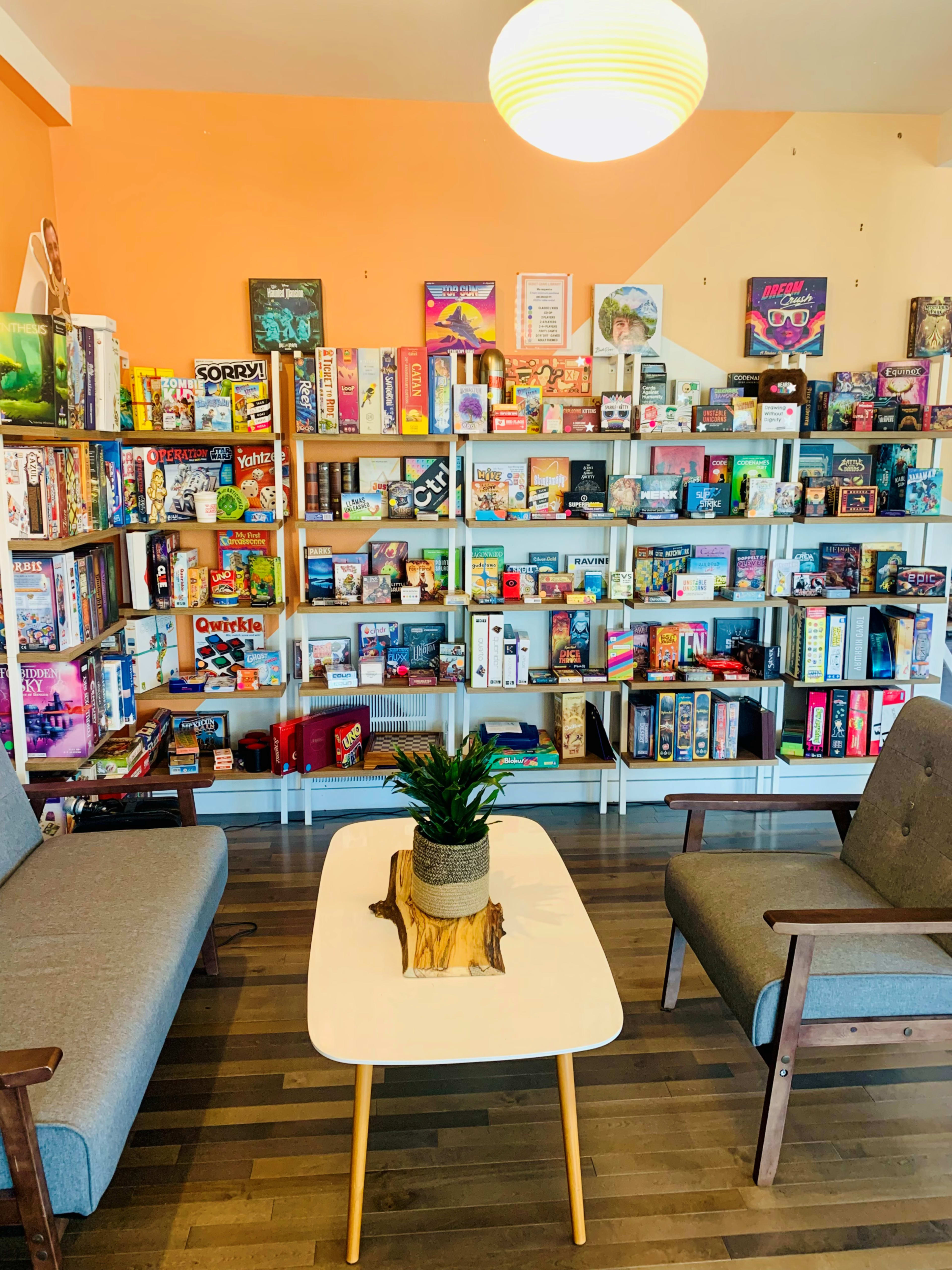 A cozy game room features two gray chairs facing a small table with a potted plant, surrounded by shelves filled with a variety of board games.