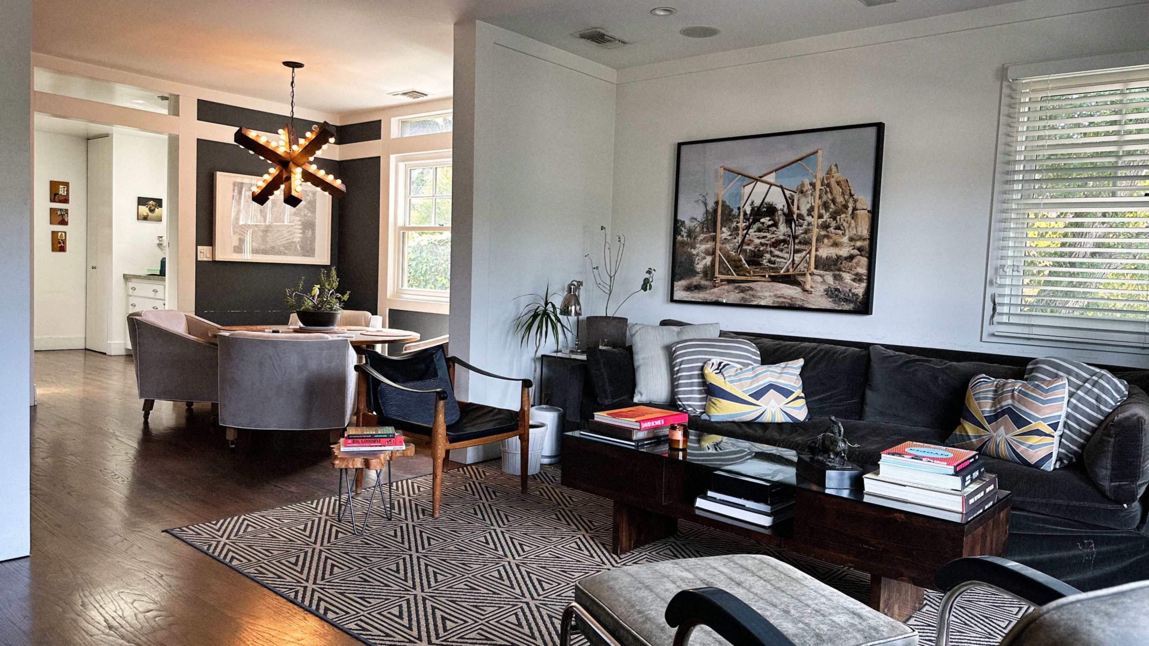 The image depicts a modern living room featuring a dark sectional sofa, a wooden coffee table stacked with books, and an art piece on the wall, with a dining area visible in the background.