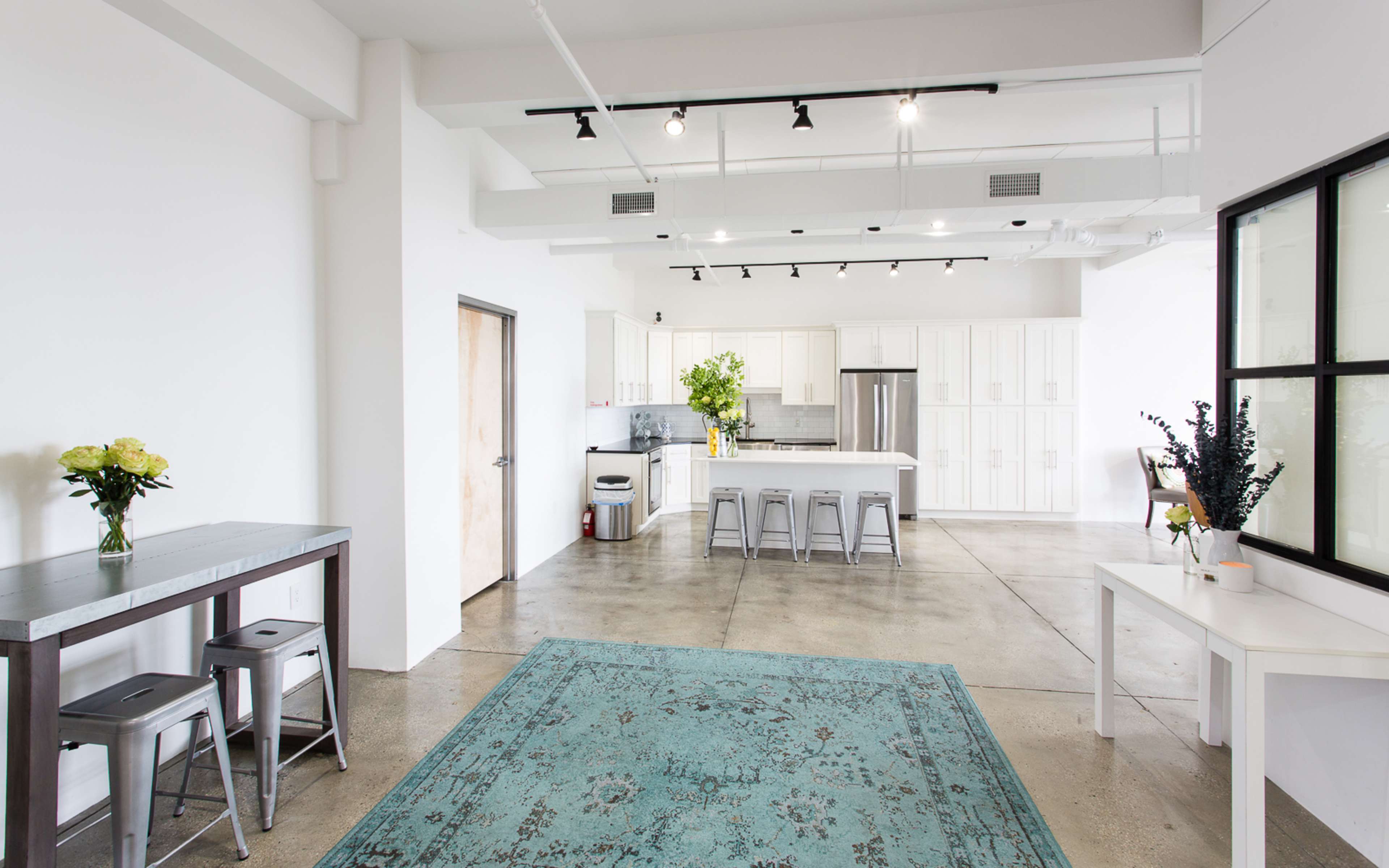 A modern kitchen area with white cabinets, a large island, and a concrete floor, complemented by a rug and flower arrangements.