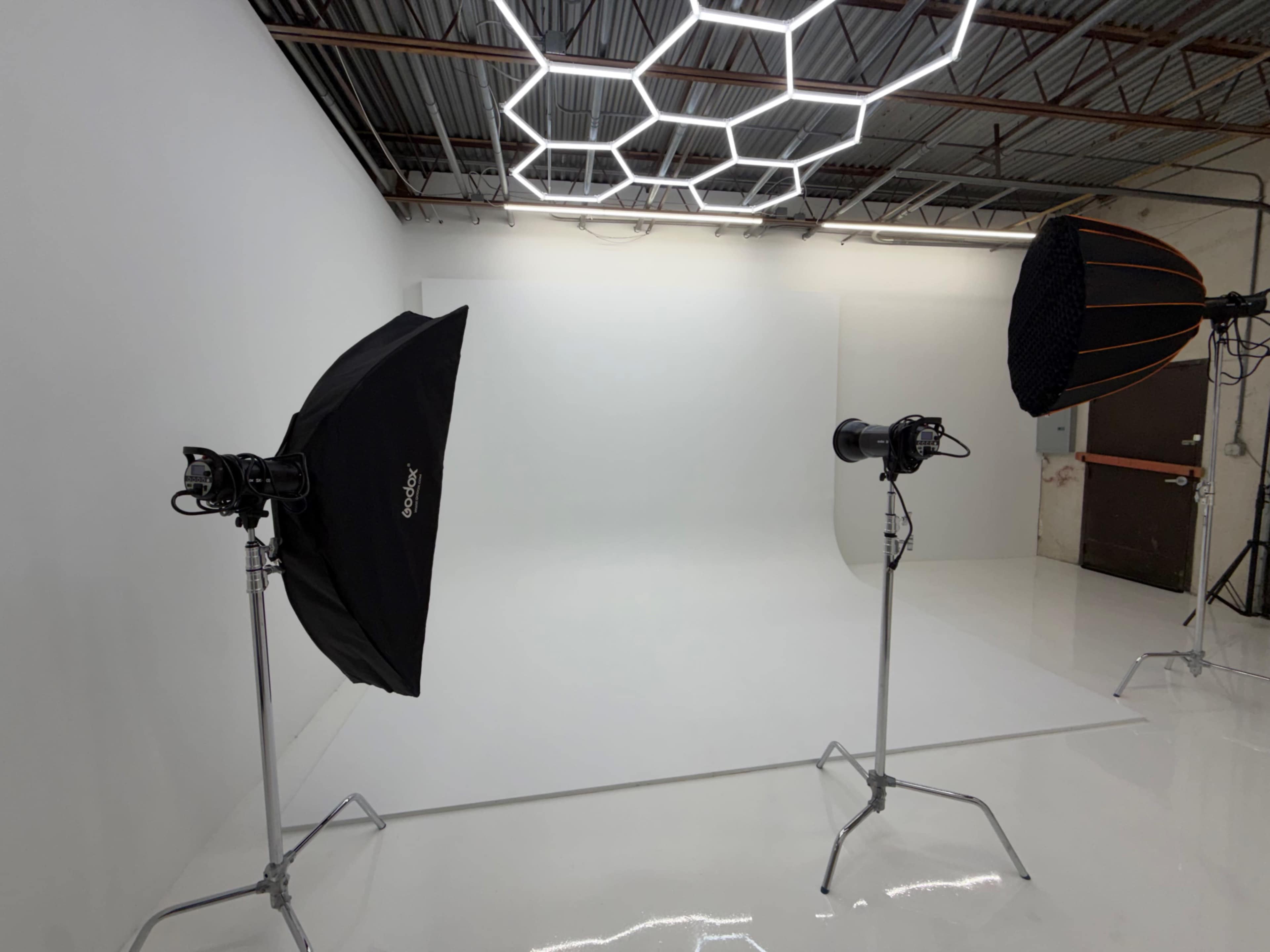 The image shows a photography studio with a seamless white backdrop, equipped with two professional lighting setups on stands.