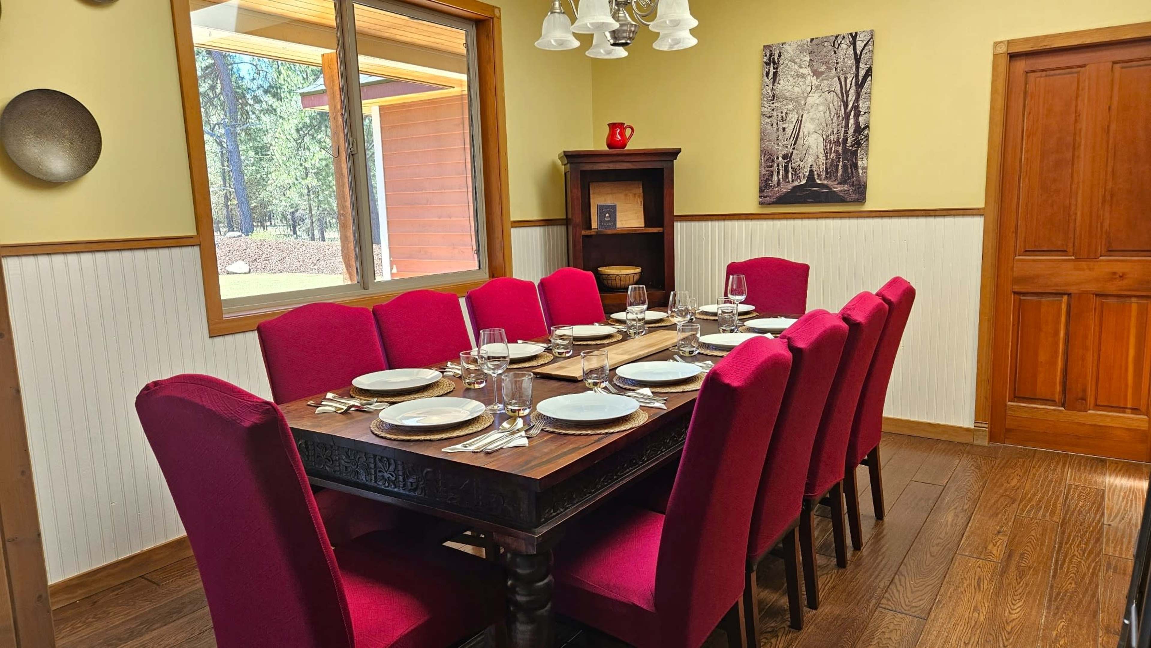 A dining room features a large wooden table set for eight, surrounded by red upholstered chairs, with a window showing a forest view and a painting on the wall.