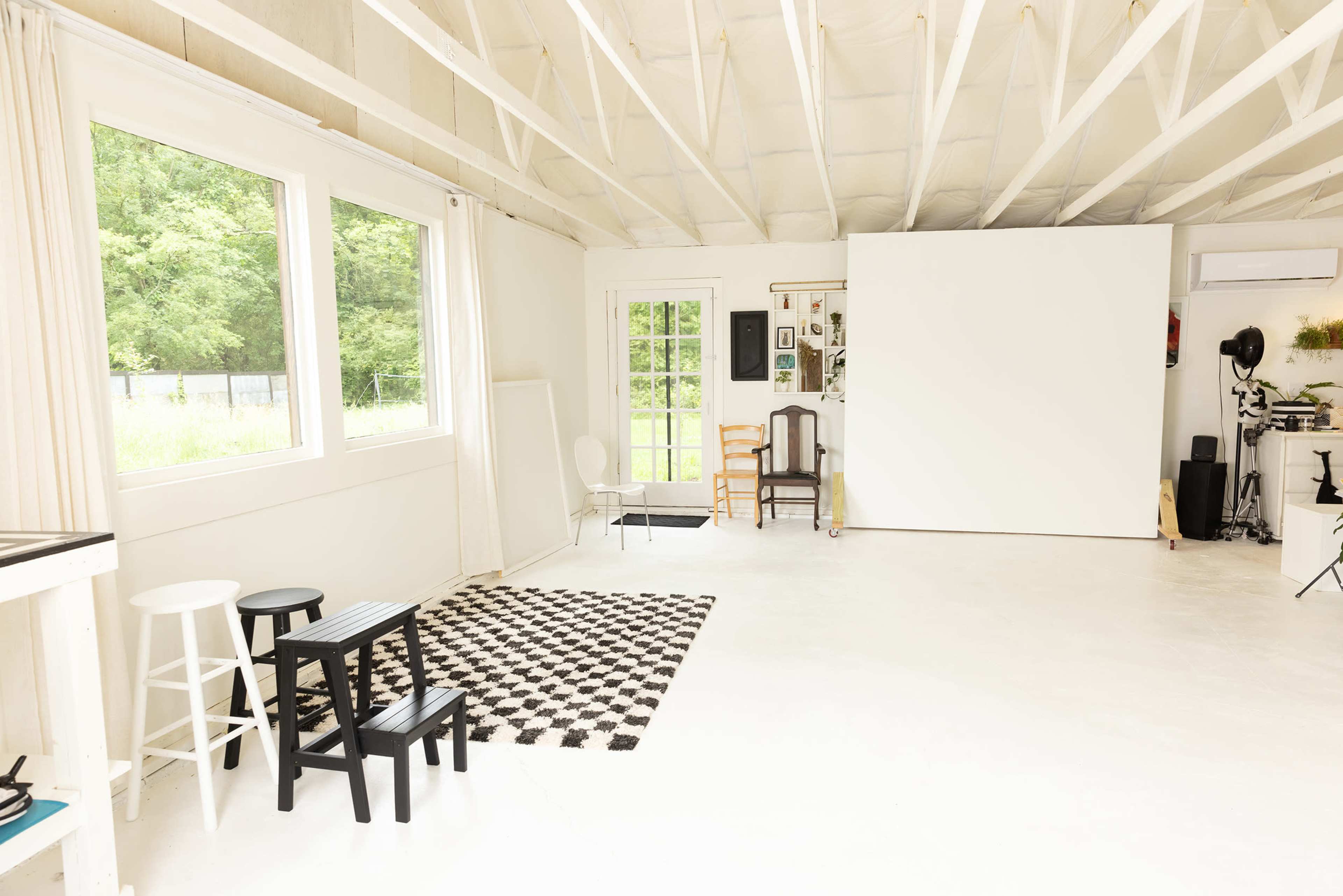 The image shows a bright, spacious room with white walls, large windows, and a large blank screen at one end, furnished with various chairs and a patterned rug.