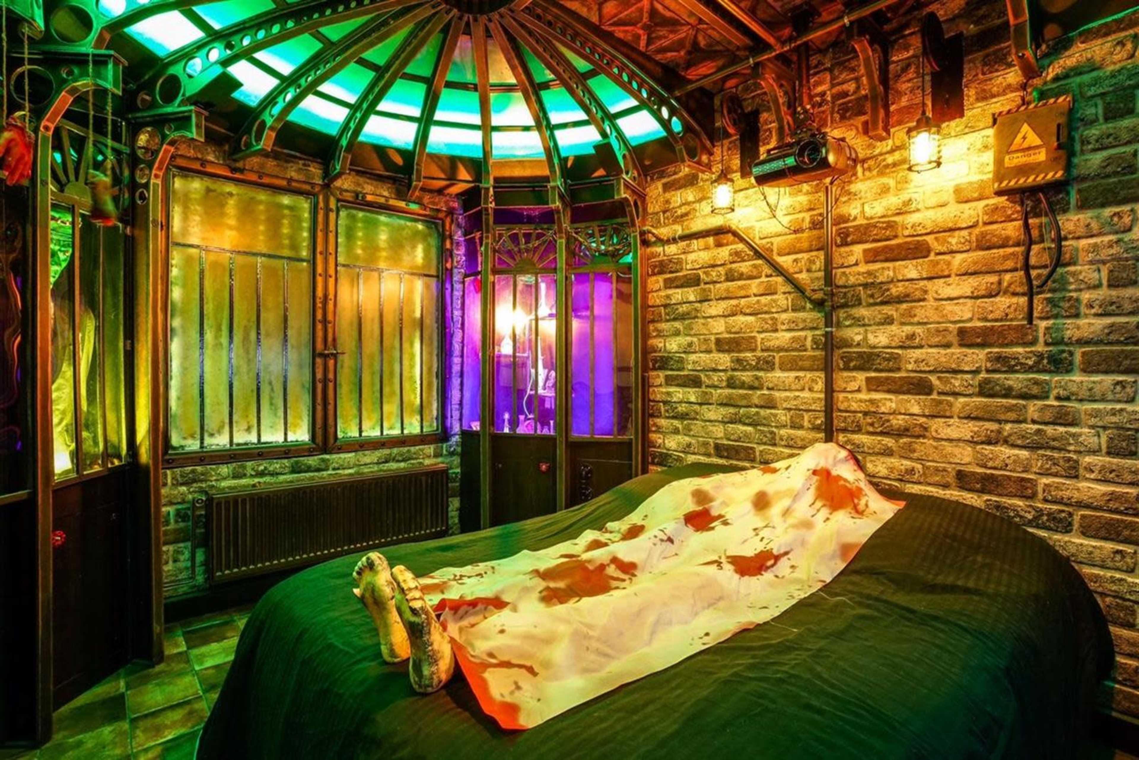 A dimly lit room features a bed with a stained sheet, surrounded by brick walls and a colorful, illuminated ceiling.