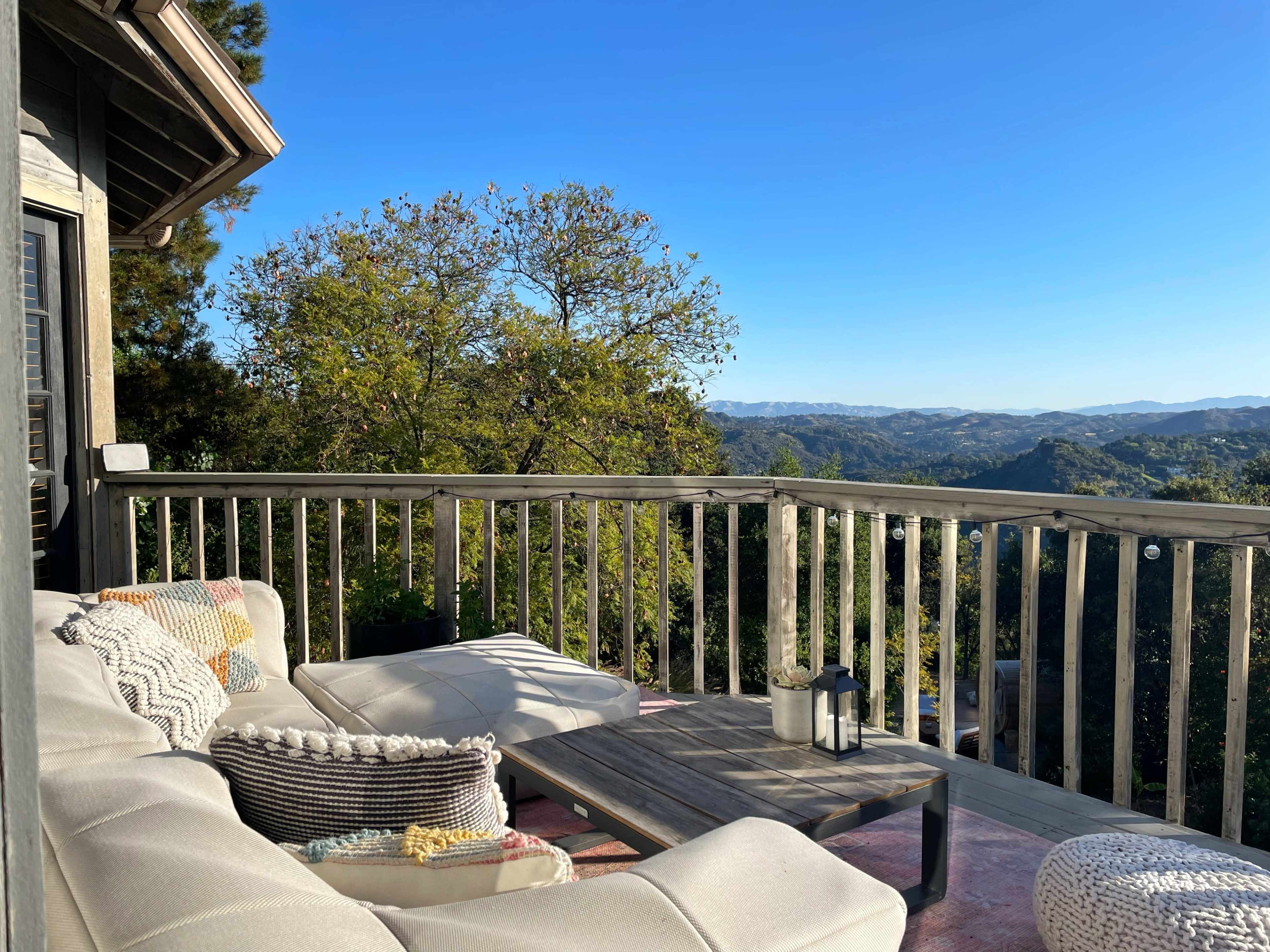 A deck with a sectional sofa and a wooden table overlooks a scenic landscape of rolling hills under a clear blue sky.