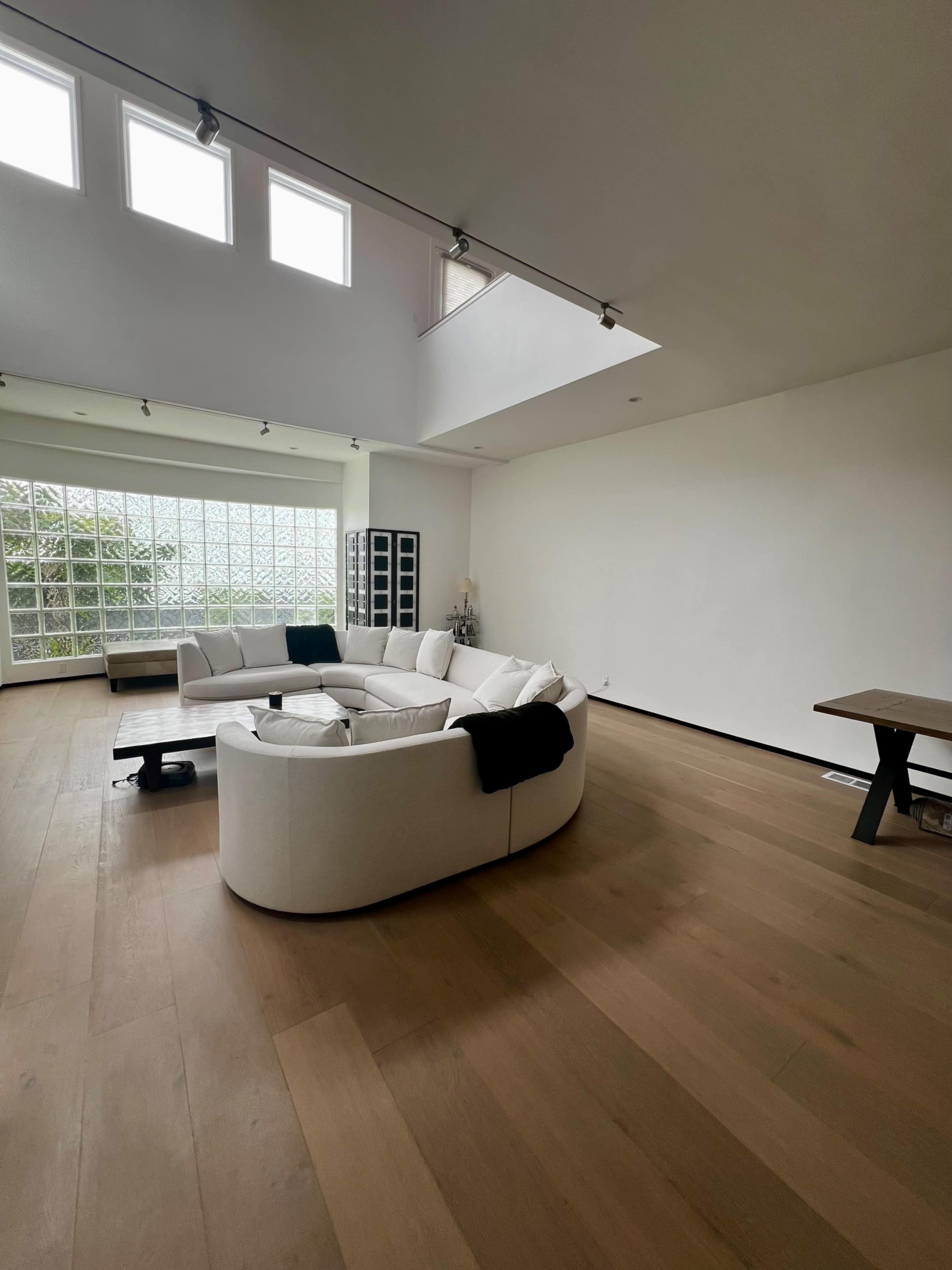 A spacious living room featuring a white sectional sofa, large windows with glass blocks, and a wooden floor.
