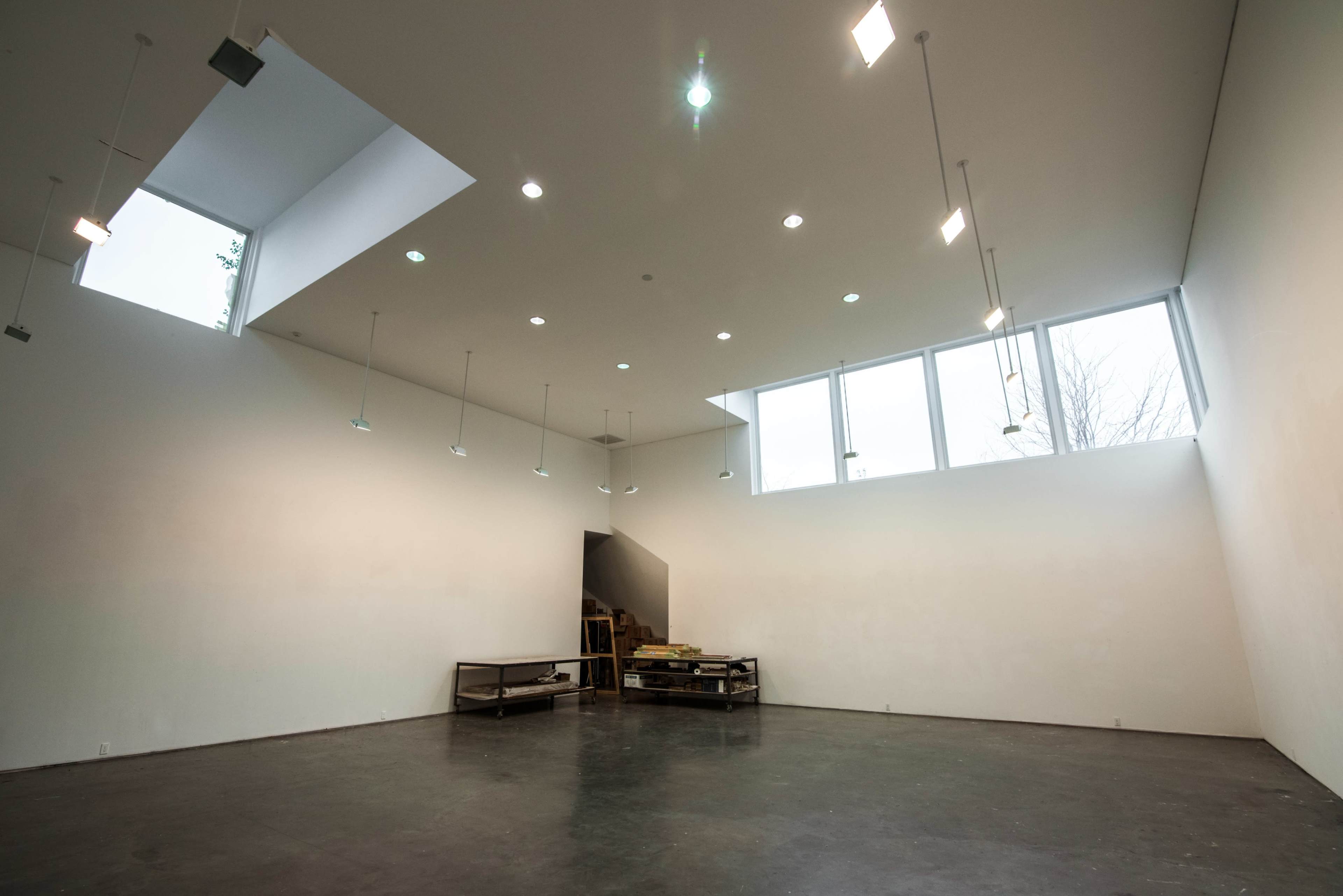 A spacious, empty gallery with large windows and overhead lighting illuminates its white walls and polished floor.