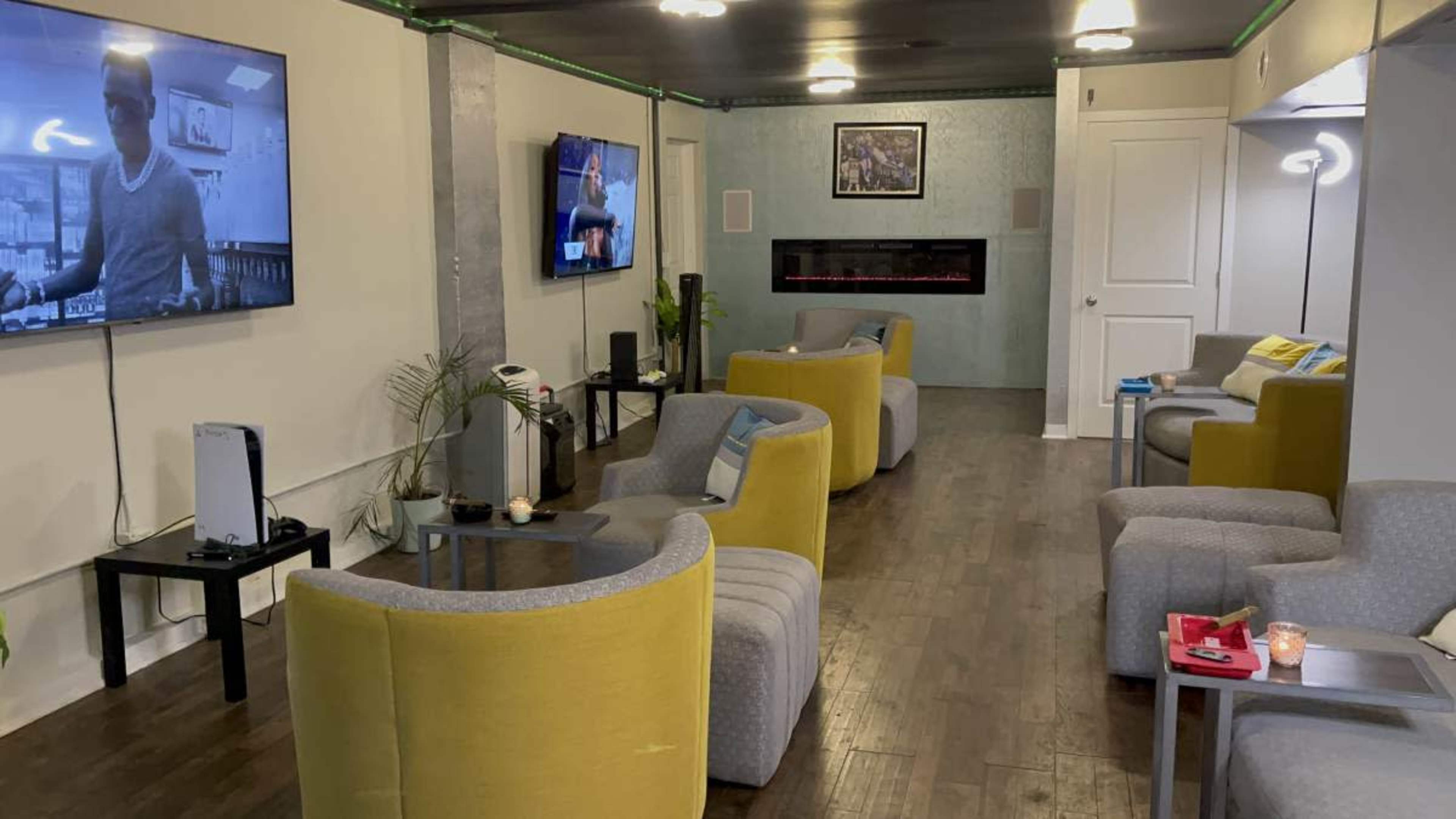 The image shows a modern lounge area with several gray and yellow armchairs arranged around small tables, featuring two televisions mounted on the walls and a decorative electric fireplace.