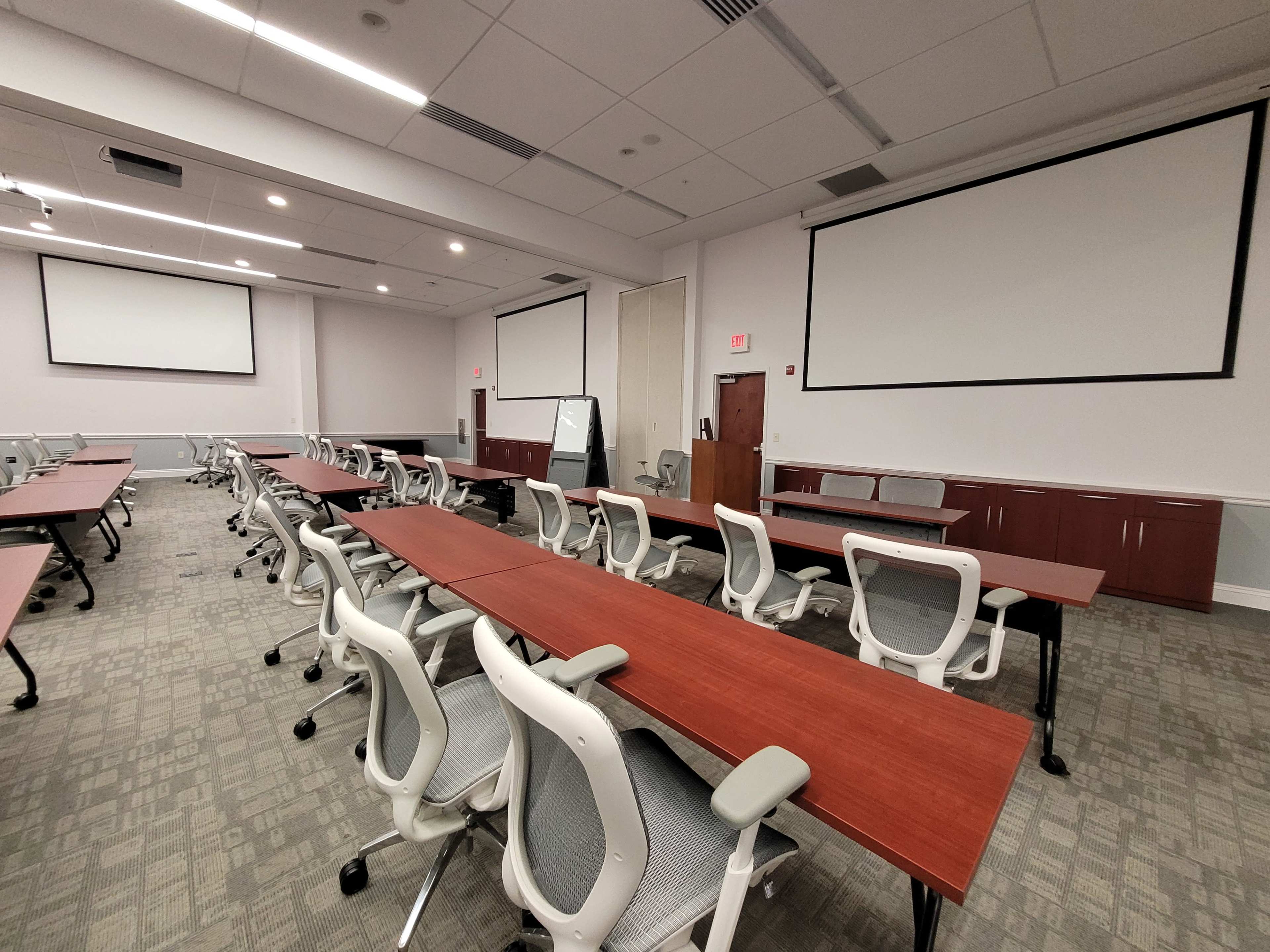 The image shows a large, empty conference room with multiple long tables and chairs arranged for a meeting, featuring dual projection screens at the front.